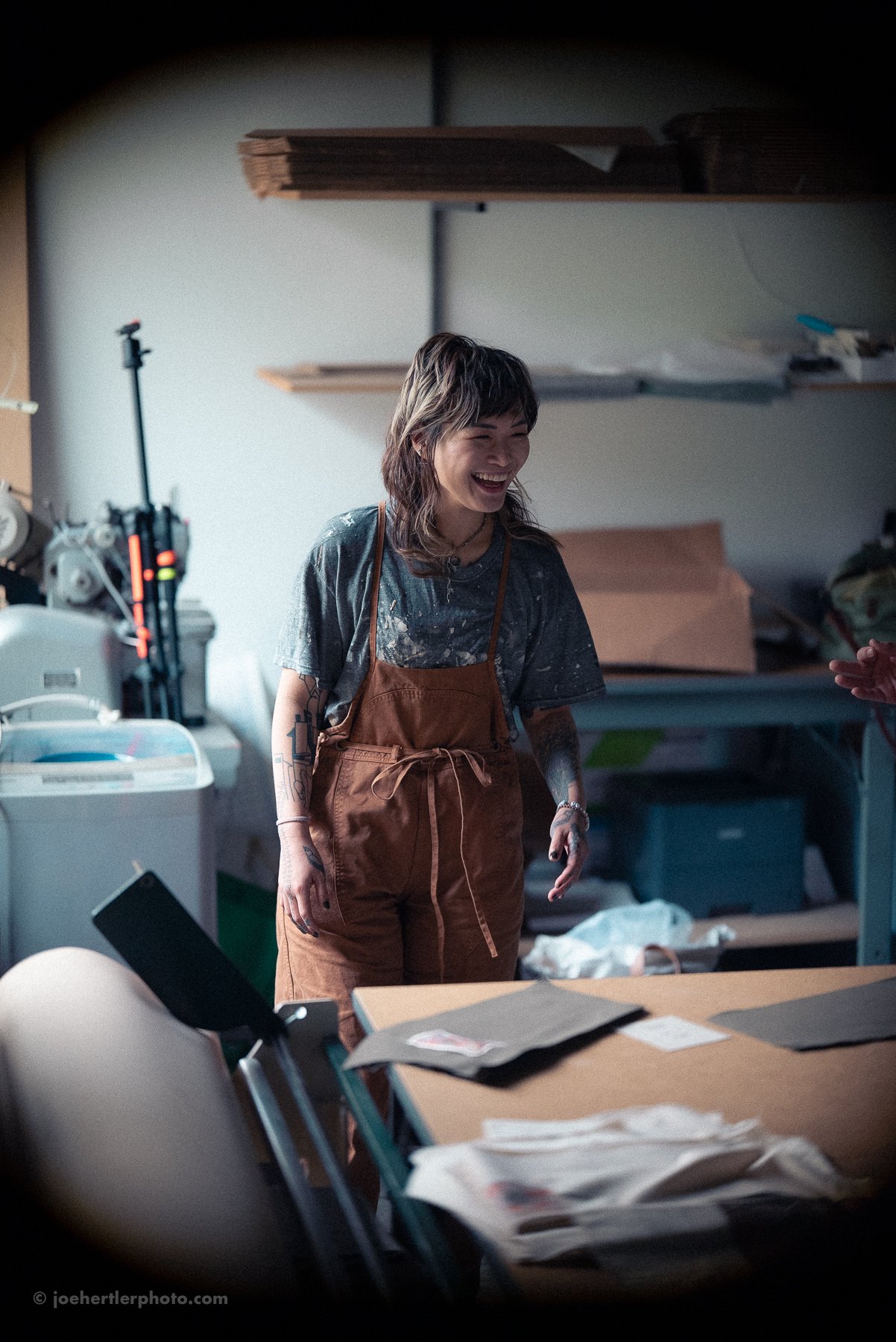 A young woman with tattoos, wearing a gray t-shirt and brown apron, is smiling and laughing in a cluttered workshop or studio with various supplies and fabrics around her.