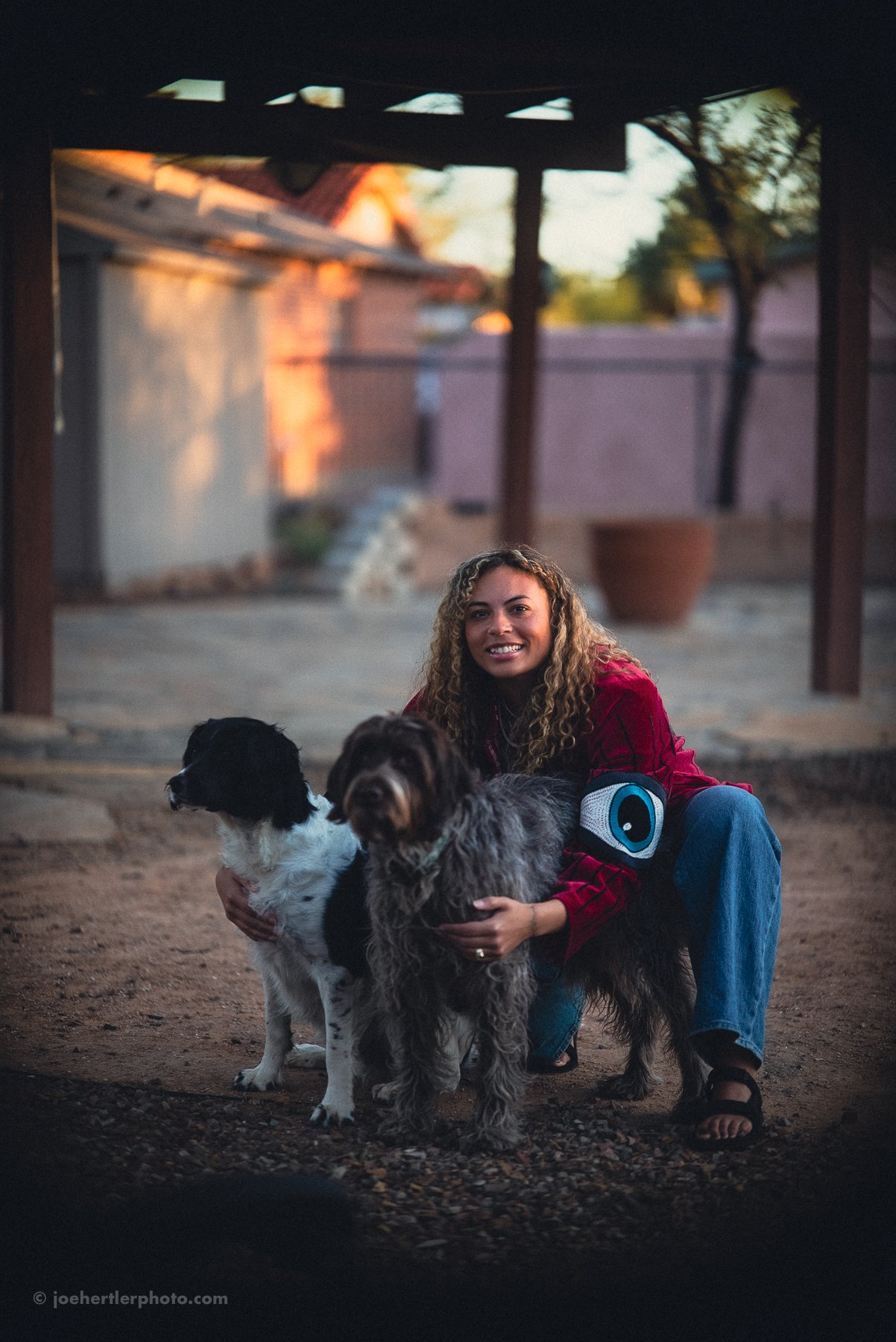 A woman crouching outdoors with two dogs, smiling at the camera during sunset.