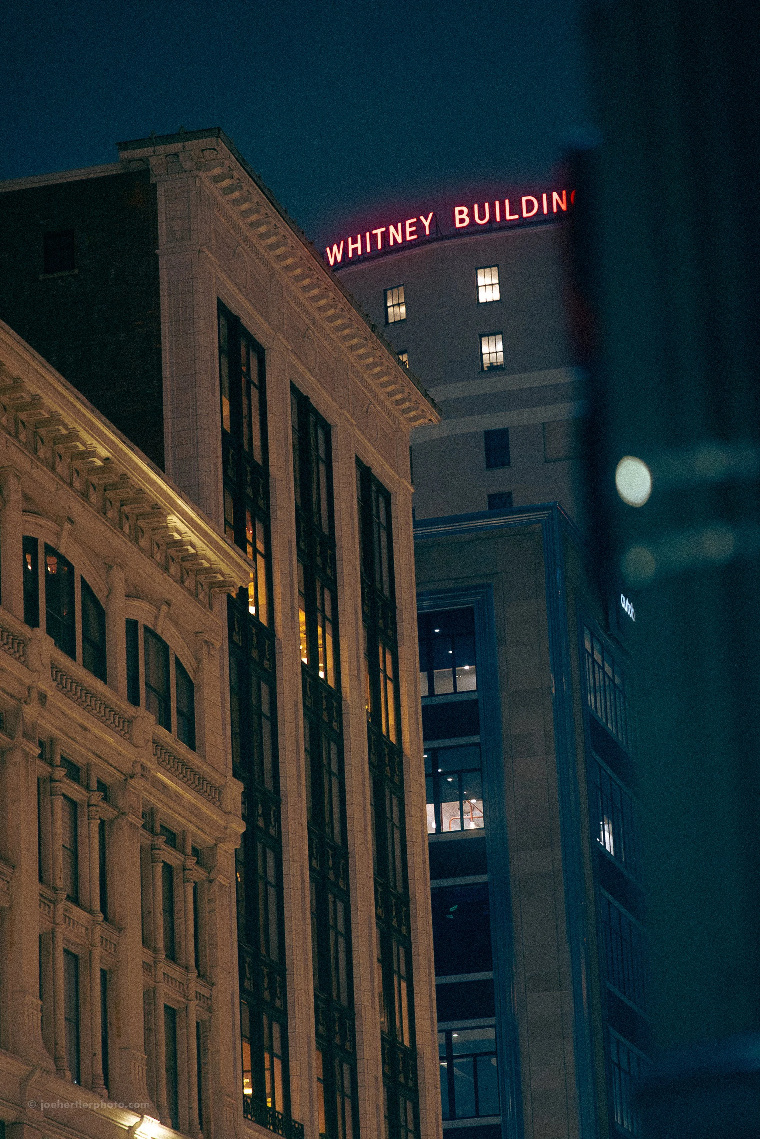 Nighttime view of tall buildings in Seattle, Washington, with the Whitney Building sign illuminated in red on top of a modern high-rise, and part of a historic building with ornate architecture in the foreground.