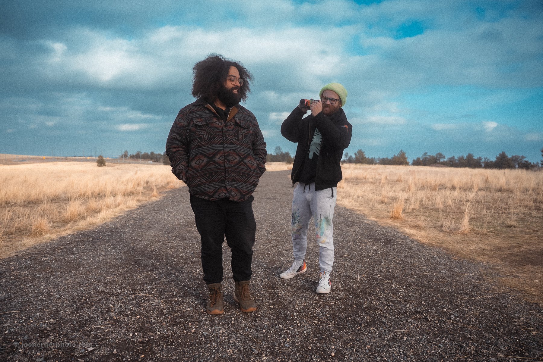 Two men standing on a gravel road in a field with dry grass, one smiling with hands in pockets, and the other holding a bottle to his ear, wearing a beanie and glasses, under a partly cloudy sky.