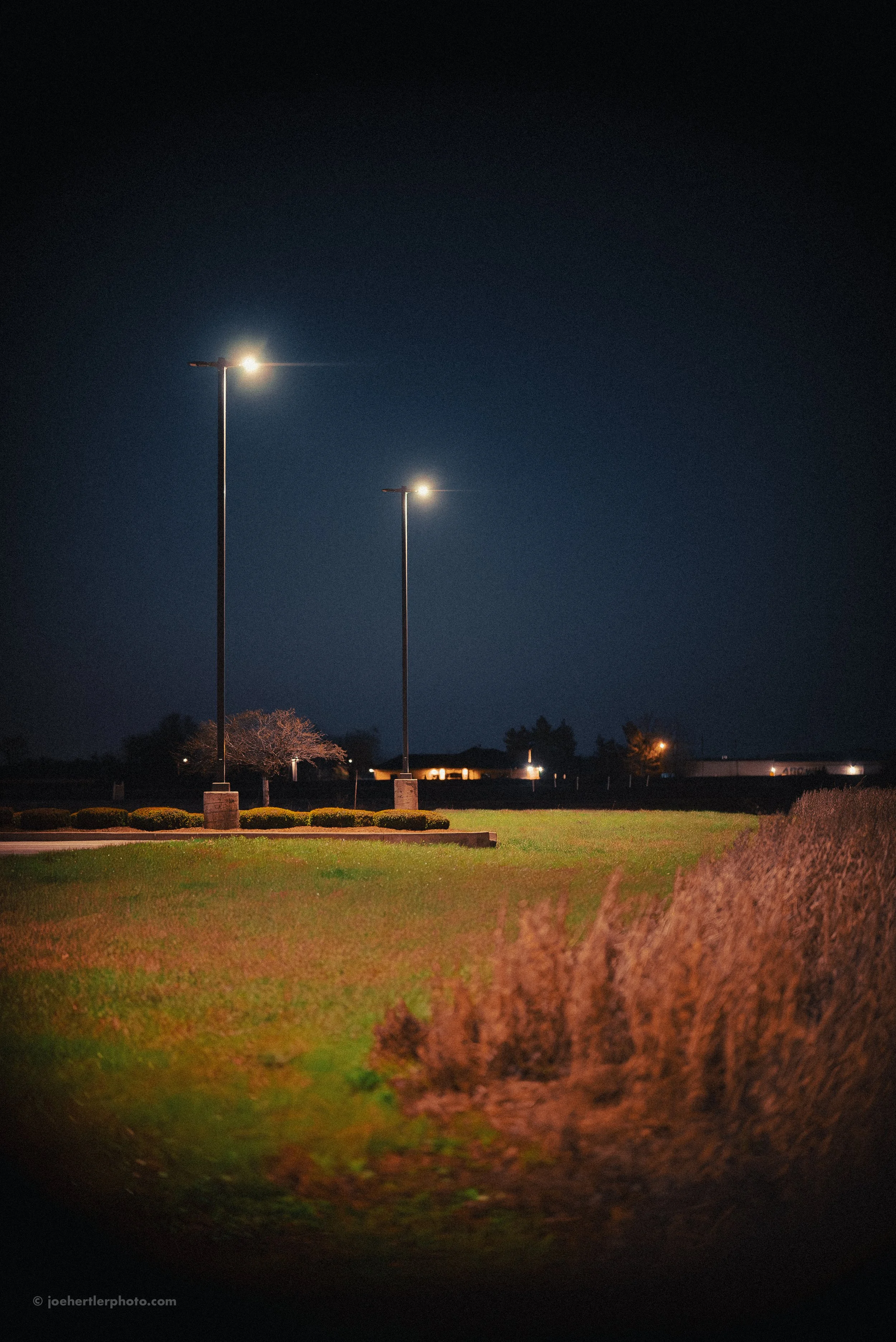 Night scene of a golf course with three tall streetlights, a grassy area, bushes, trees in the background, and a dark sky.