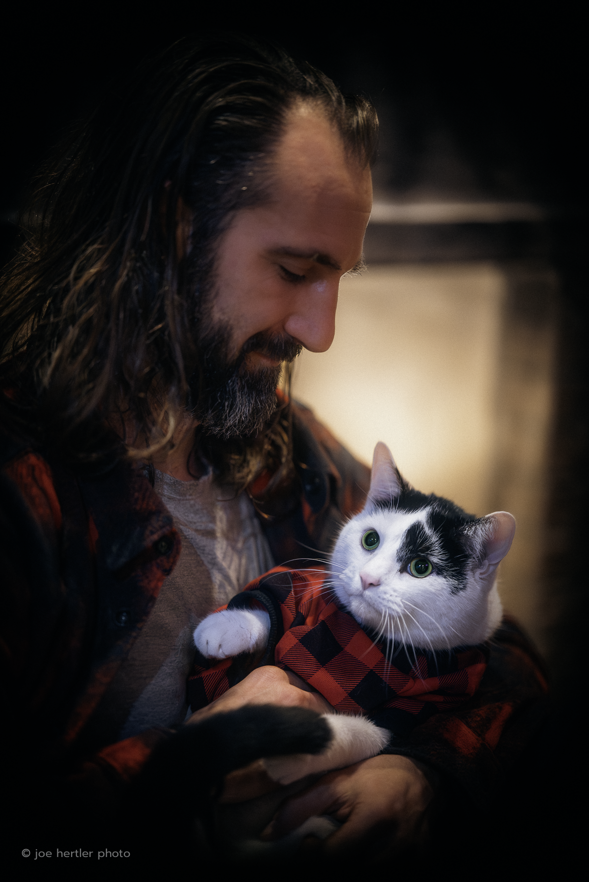 A man with long hair and a beard looks affectionately at a black and white cat wearing a red and black plaid jacket, the man holding the cat in his arms indoors.