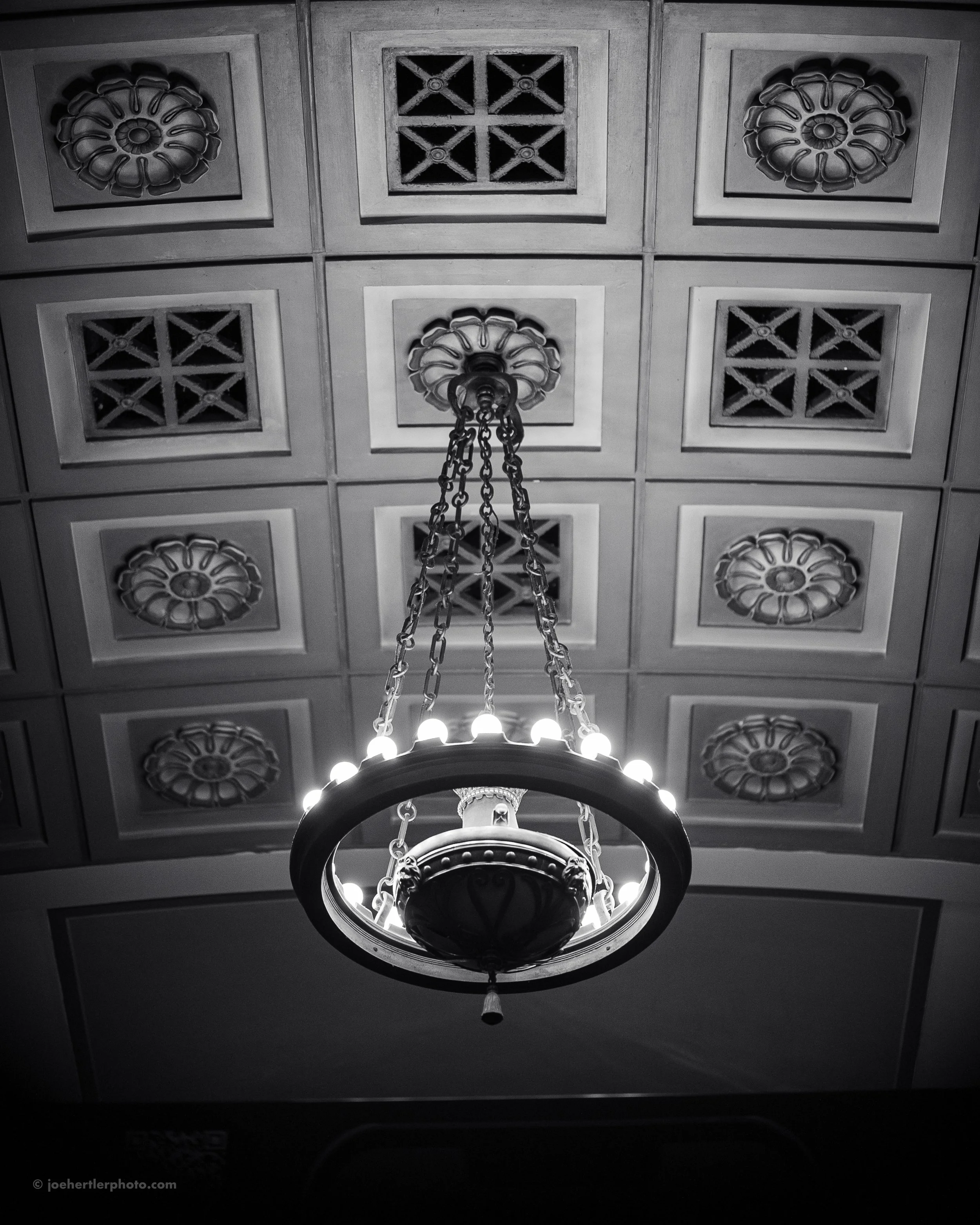 Black and white photo of ornate ceiling with a hanging chandelier with chained rings and decorative ceiling tiles.