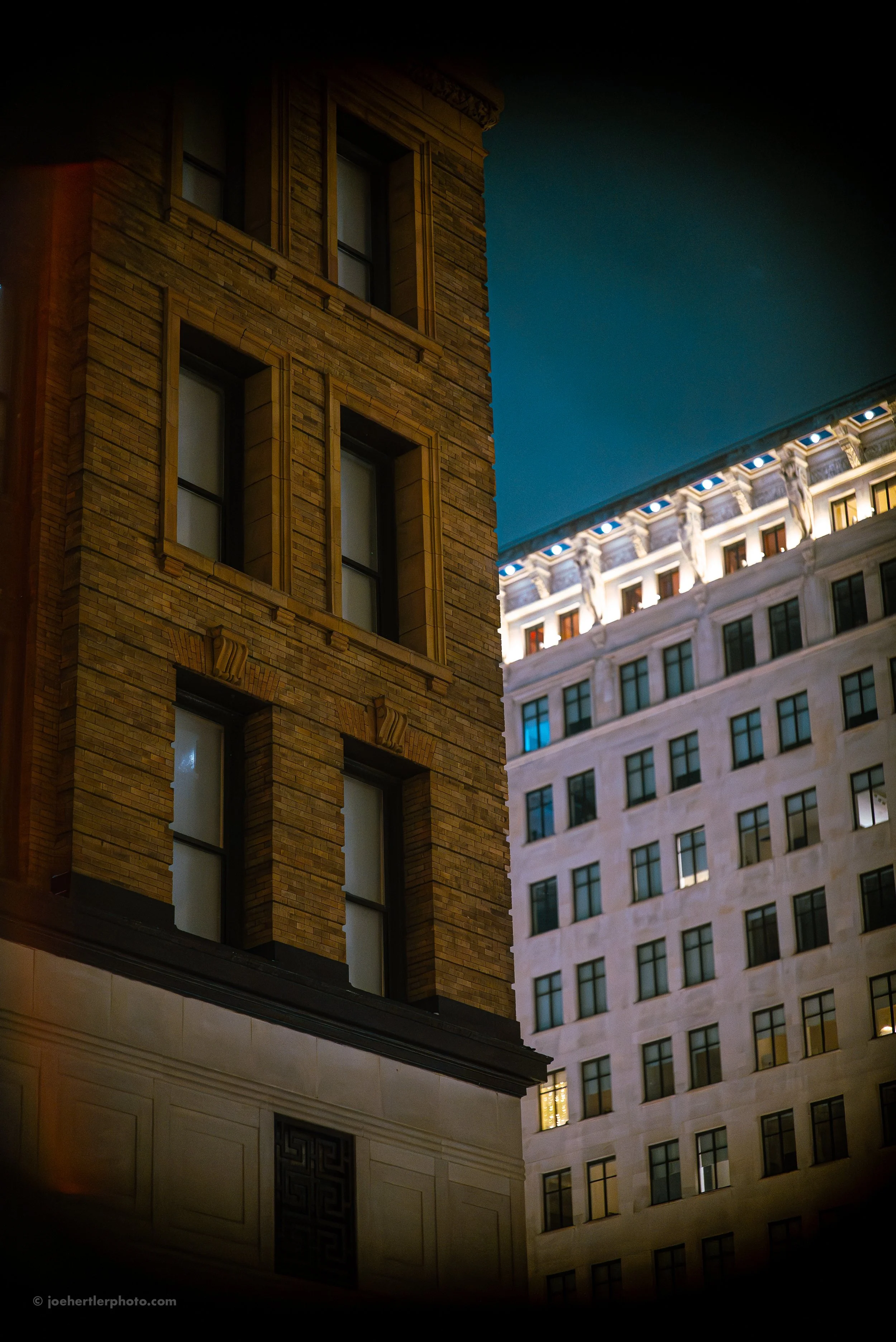 Nighttime view of two tall buildings with illuminated windows and architectural details in an urban setting.
