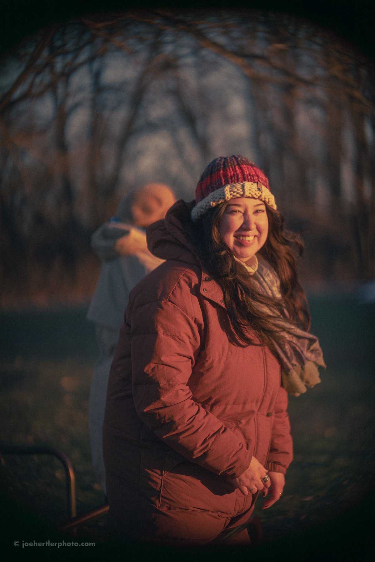 A woman smiling and wearing a knit hat and a winter jacket outdoors during dusk or night, with trees in the background.