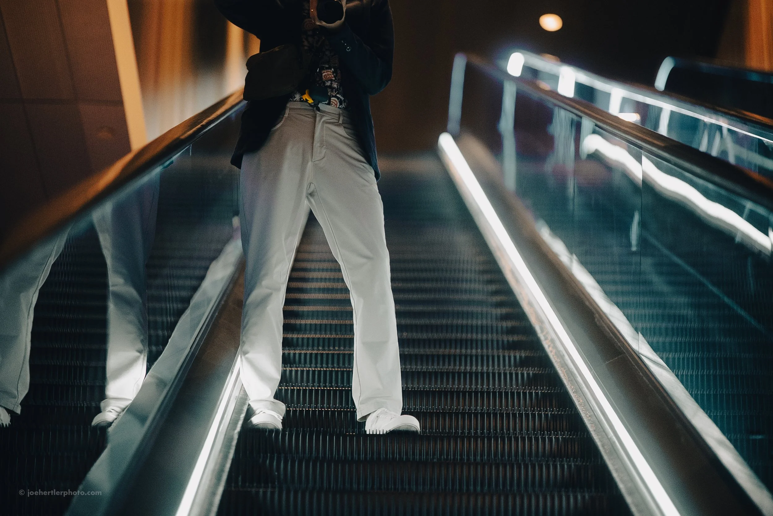 Person standing on an escalator, wearing beige pants and a dark jacket, with a reflection visible on the escalator's glass side.