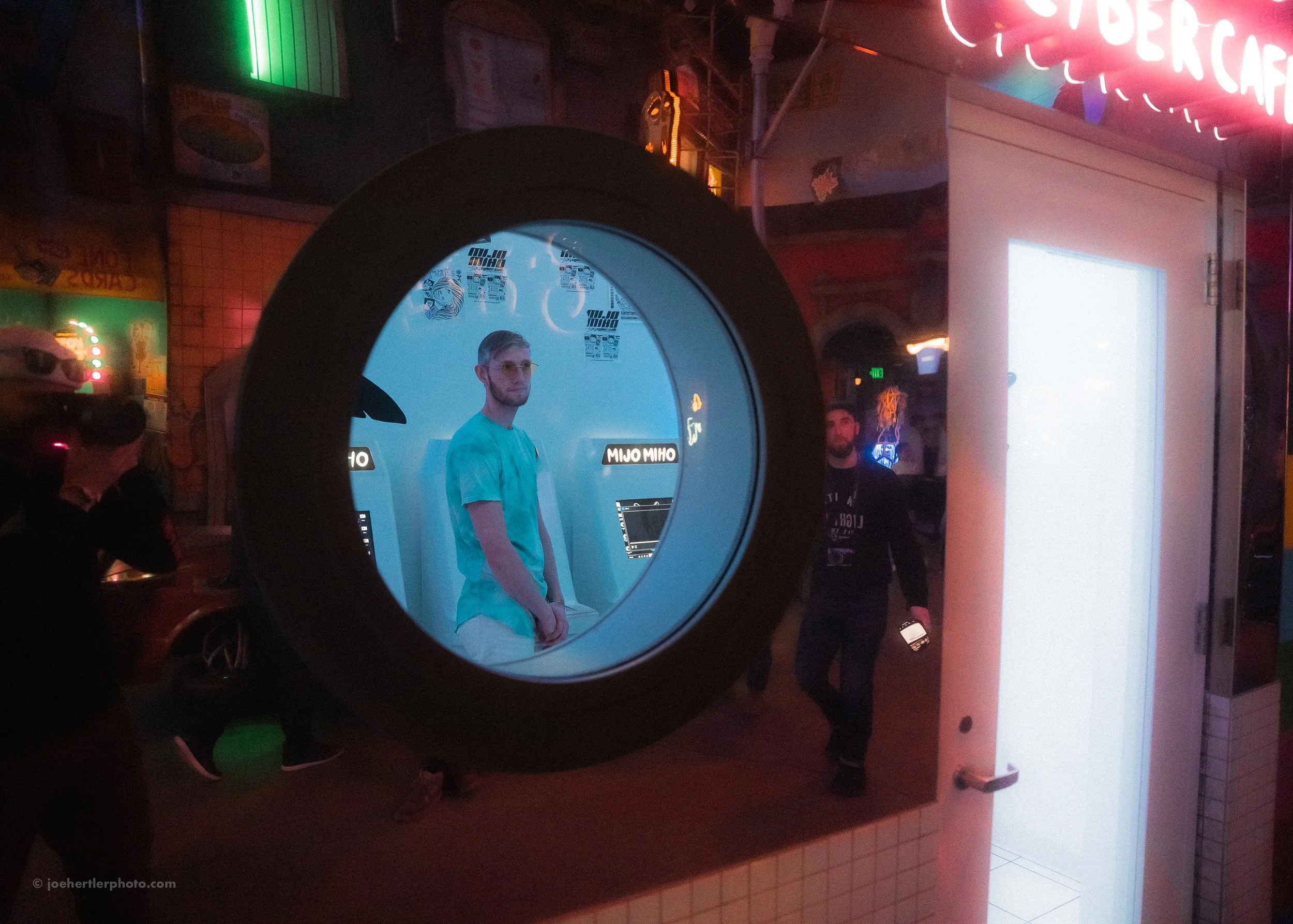 A man in a turquoise shirt is inside a circular photo booth with signs that say 'MIJO MIJO.' The booth has a blue interior. The photo is taken through the circular opening of the booth. There are other people and neon signs in the background.