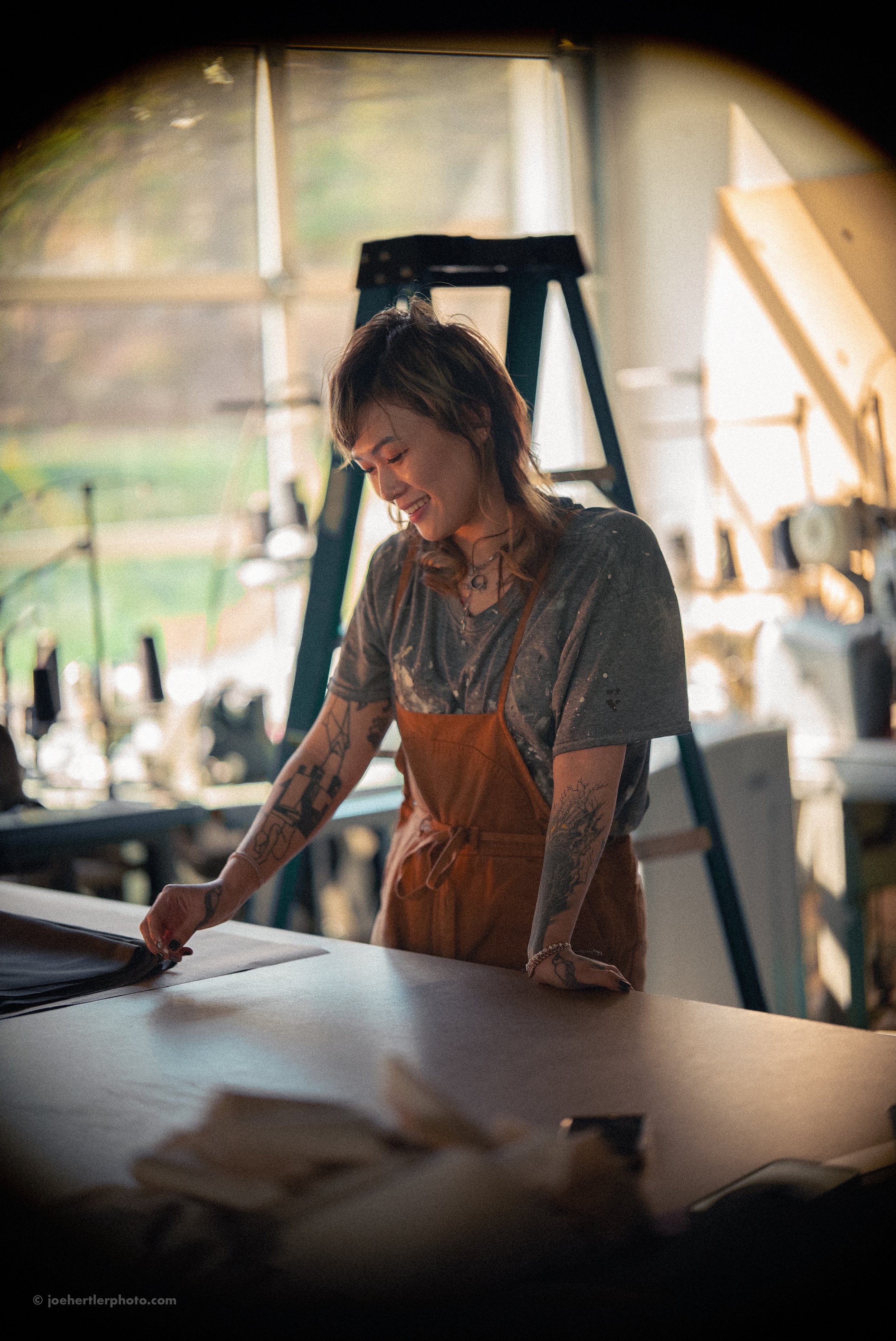 A smiling woman with tattoos and wavy hair wearing an apron, working at a craft or art studio with natural light.