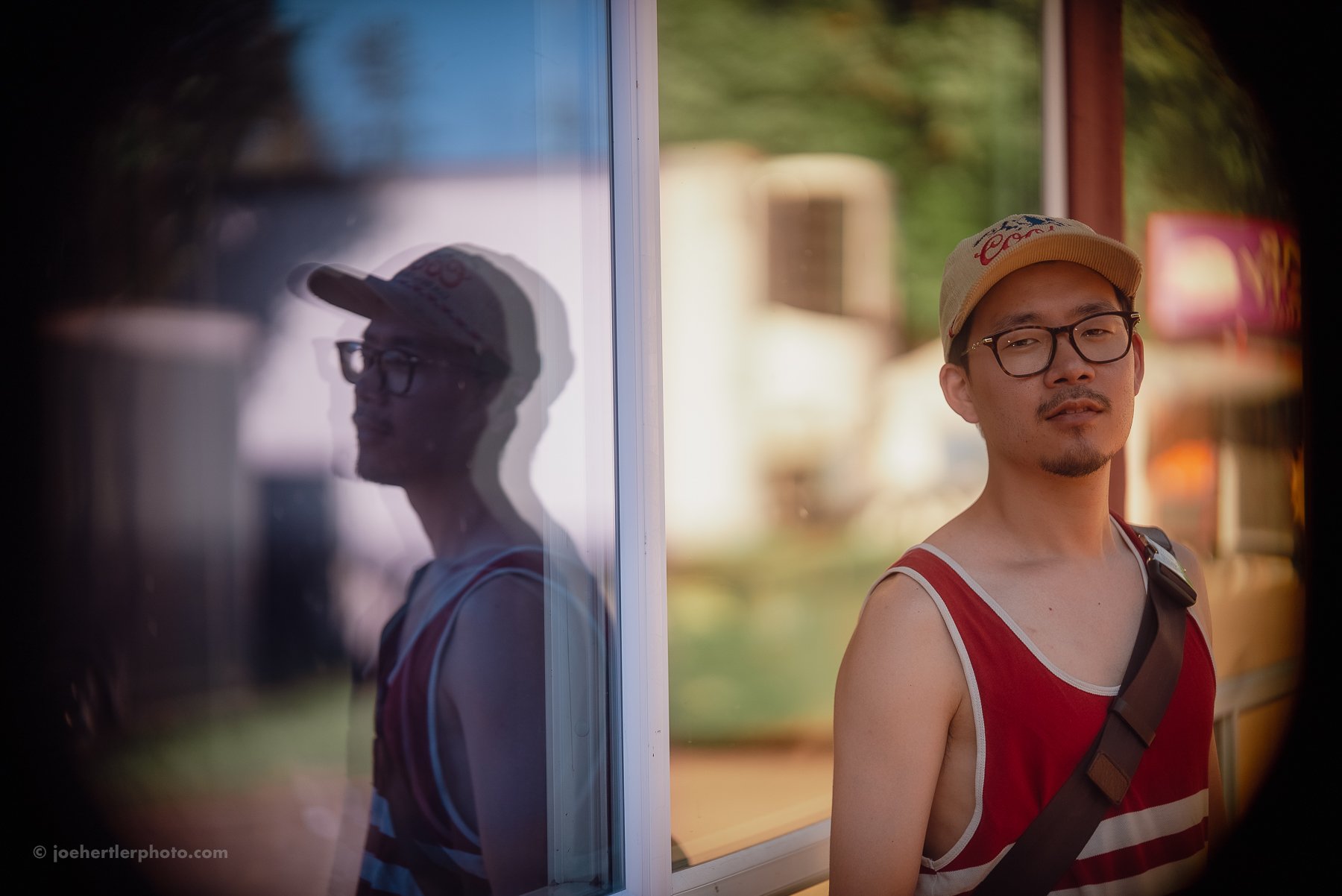 A young man in a red and white striped tank top and beige baseball cap winks at the camera during daytime, standing next to a reflective glass window that shows his shadow and reflects a blurry outdoor scene.