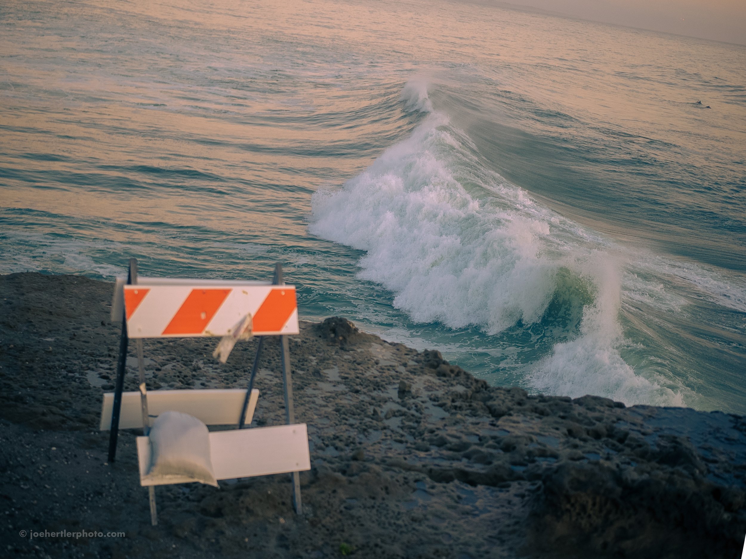 A construction barrier on a rocky shoreline facing the ocean with waves crashing and a small boat near a white surf.
