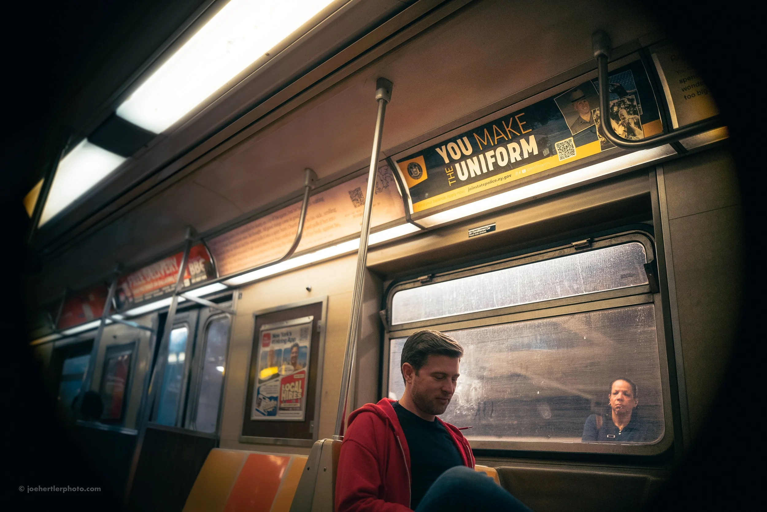 A man in a red hoodie sitting on a subway train next to a window, with a woman outside looking in reflection visible in the window.
