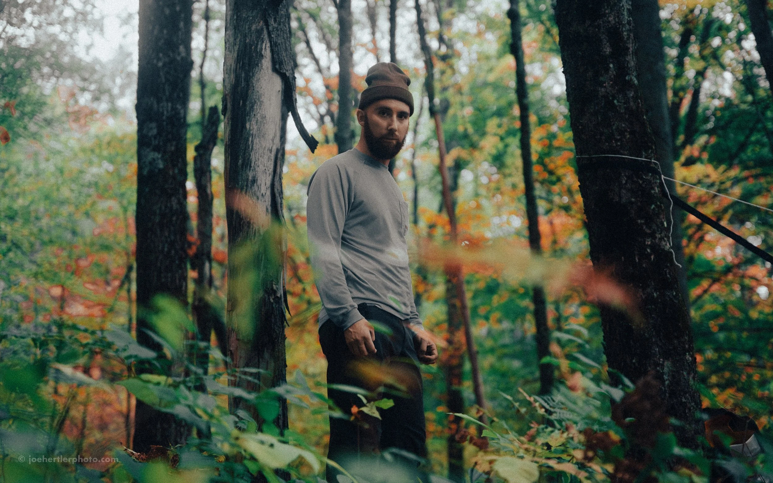 A man stands in a forest, surrounded by trees with autumn foliage, wearing a beanie and casual clothing, looking towards the camera.