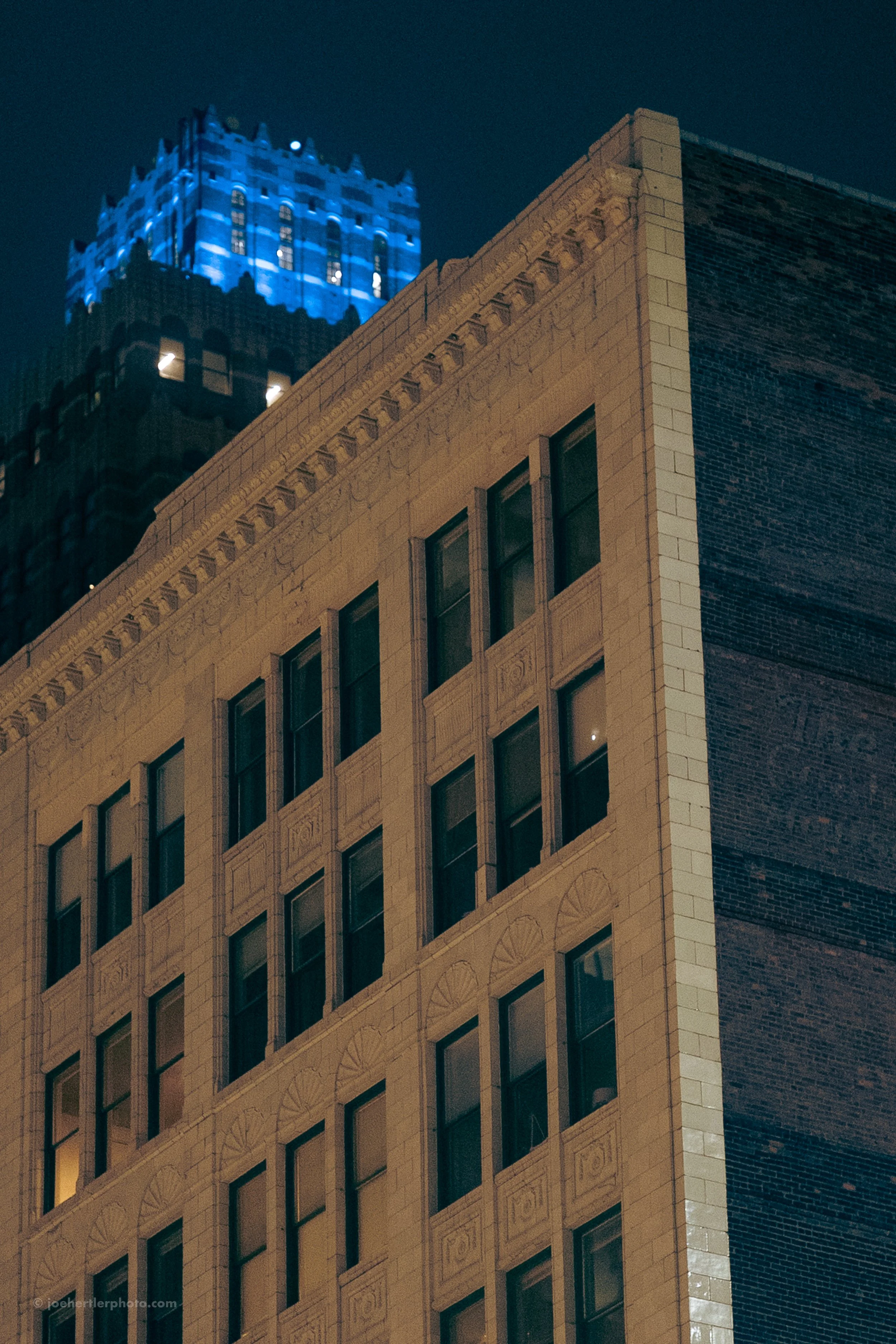 Nighttime view of buildings in a city, with the top of a large beige building featuring ornate window frames and a dark brick building on the side, and a tall skyscraper in the background illuminated with blue lights.