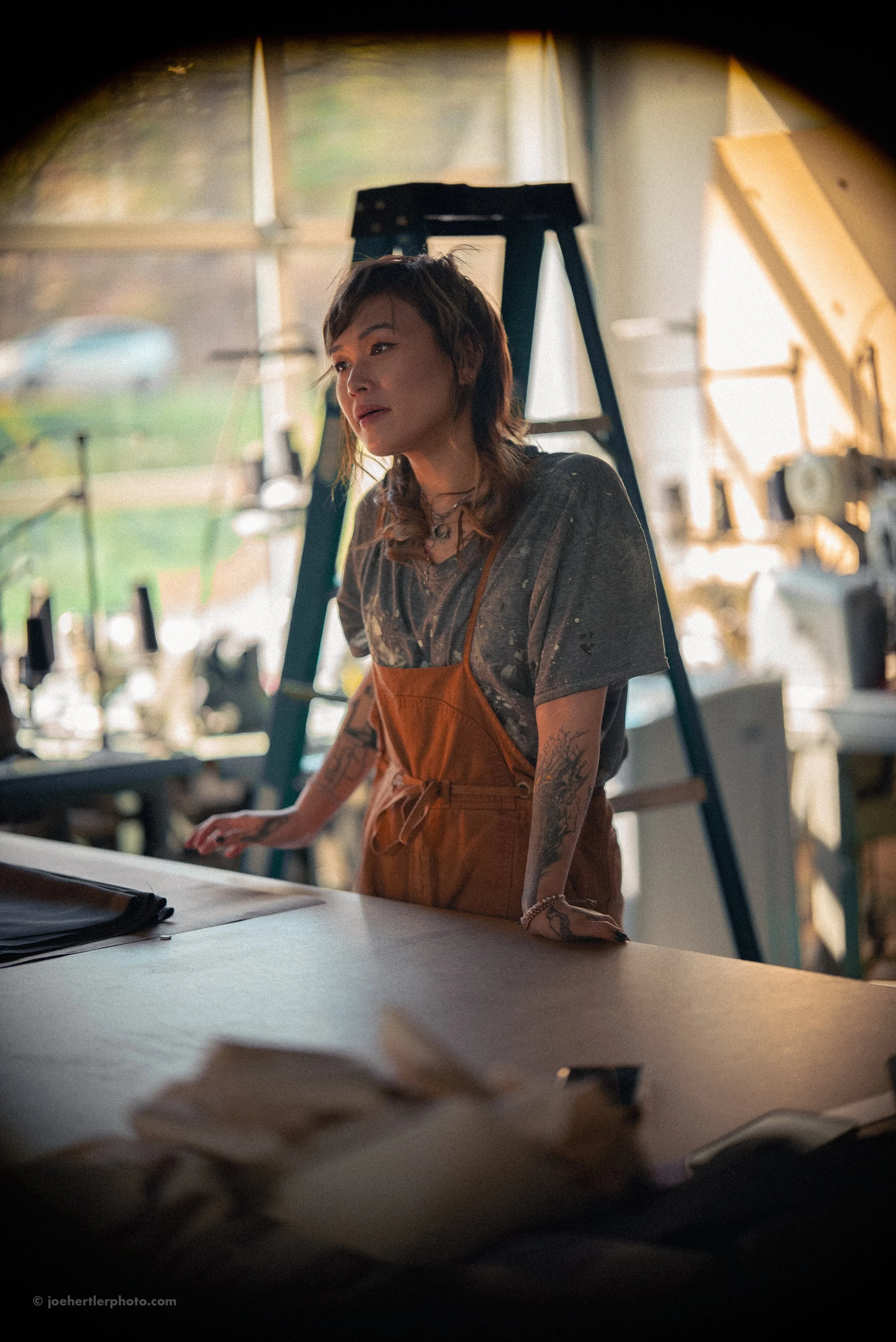 A woman with tattoos and layered hair in a workshop, wearing an orange apron and looking to the side, with tools and equipment in the background.