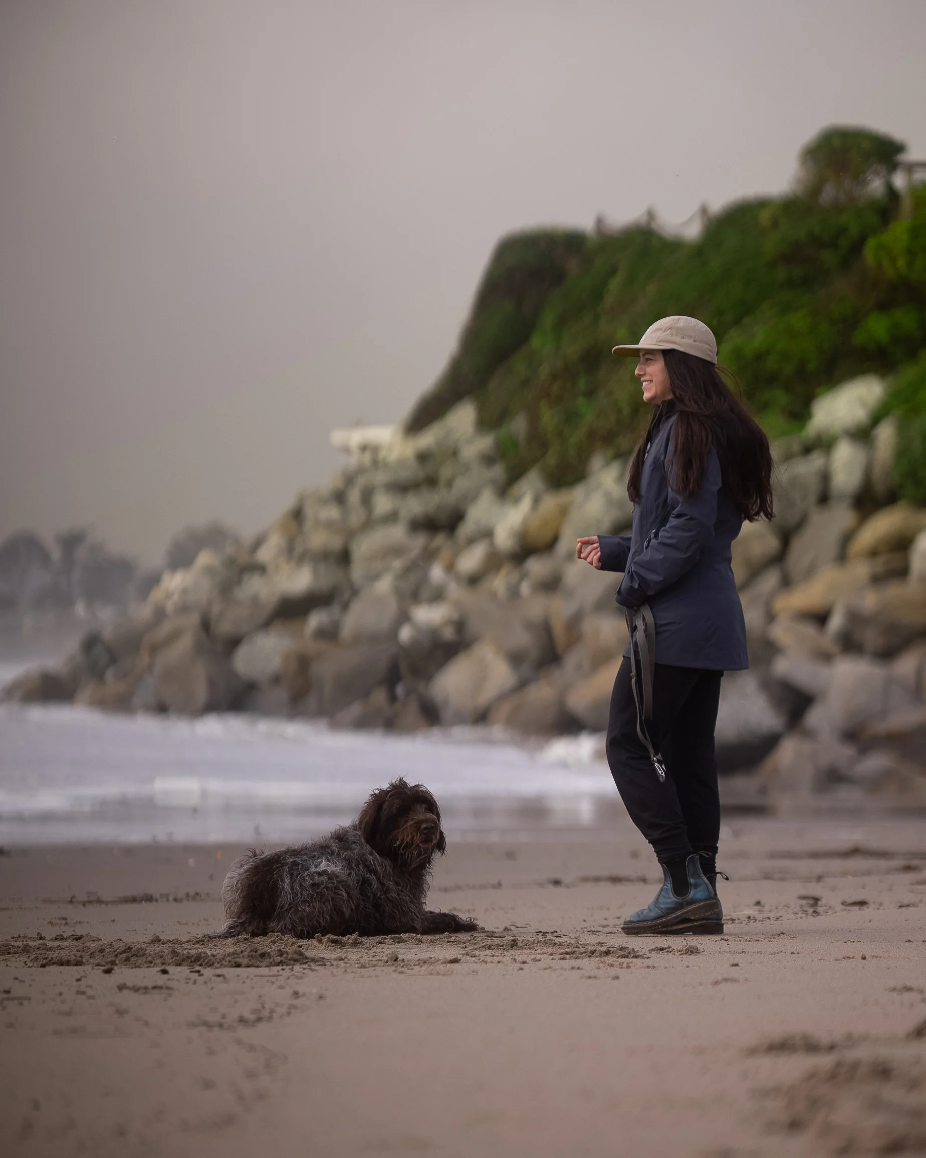 A woman in a beige hat and dark jacket stands on a sandy beach next to a small, scruffy brown dog, with rocky cliffs and greenery in the background.