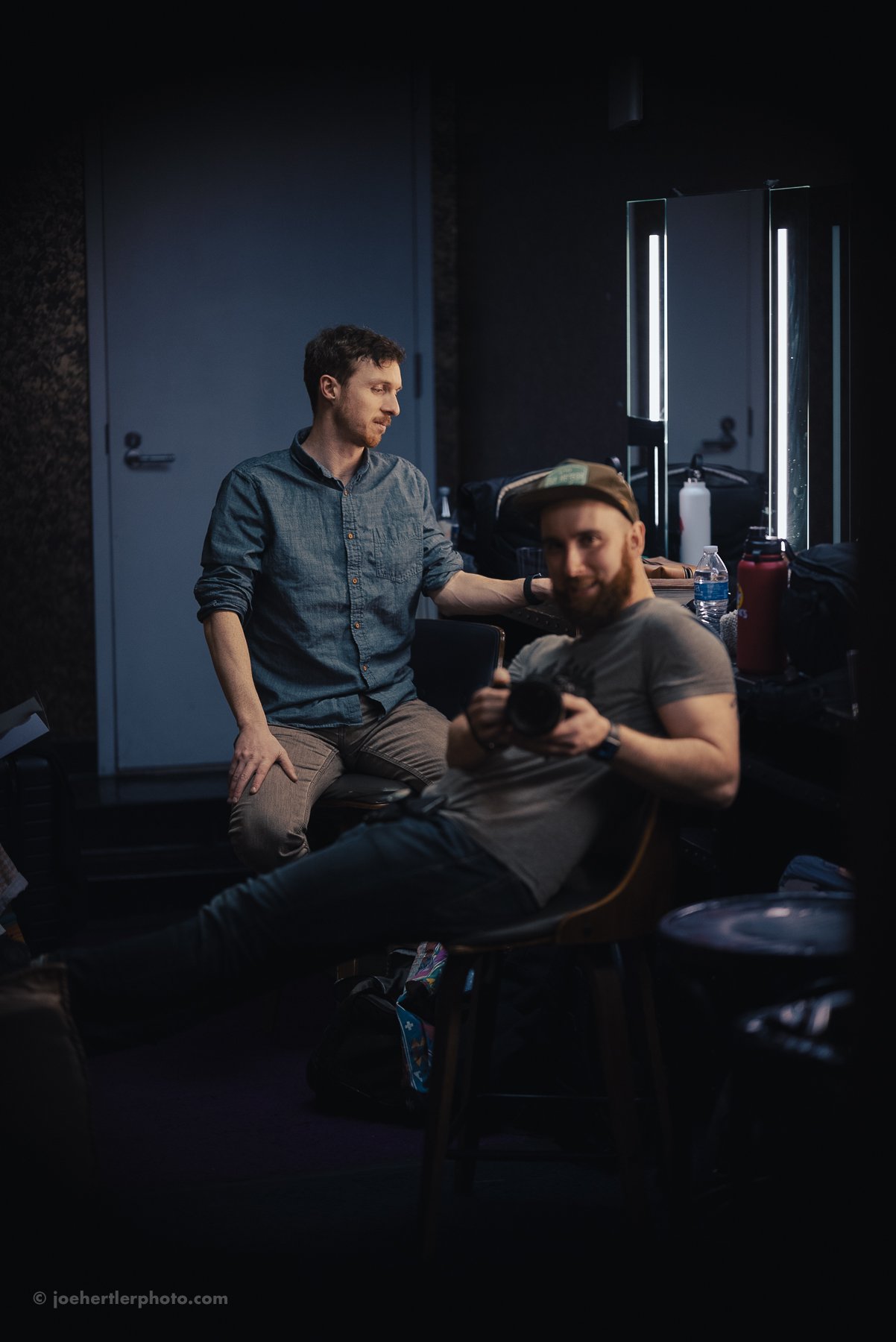 Two men in a dimly lit room with mirrors and illuminated vertical lights. One man is sitting on a chair holding a camera and smiling, while the other is standing beside him, resting his hand on the seated man's shoulder and looking down.