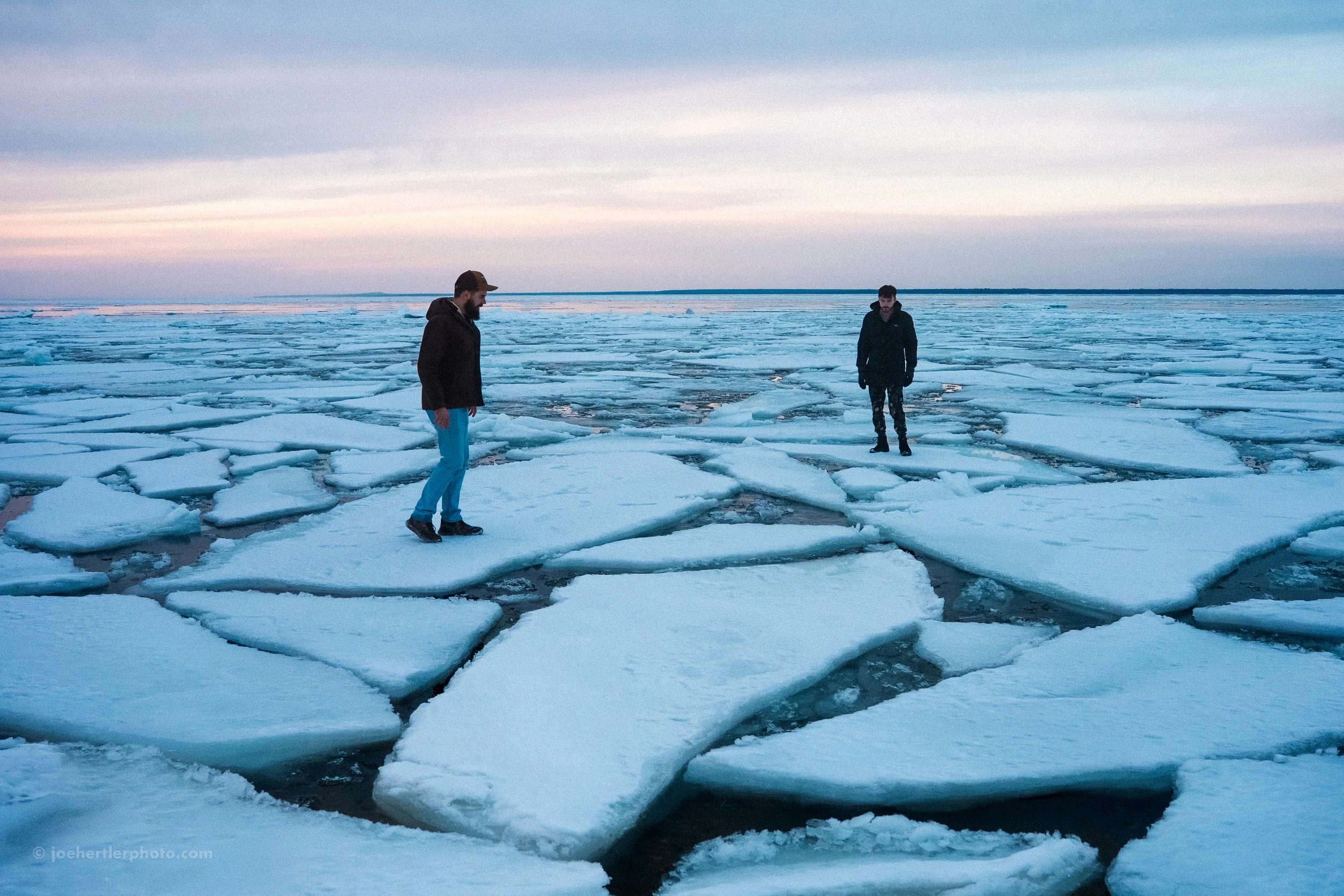 Two men walking on floating ice sheets in a frozen body of water at dusk.