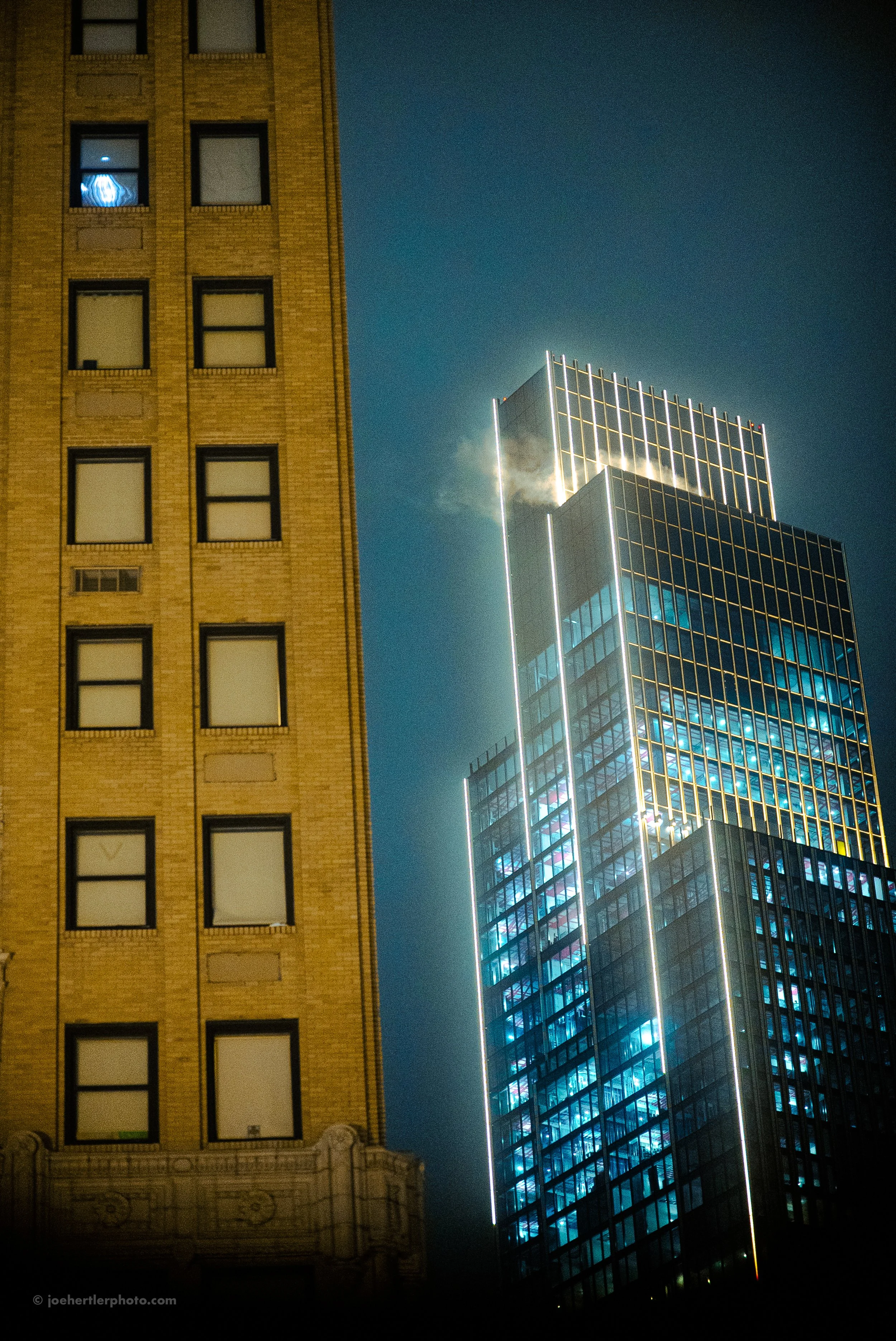 Night view of two contrasting buildings in a city, one old with a brick facade and windows, the other a modern glass high-rise with illuminated edges and a plume of smoke or vapor near the top.