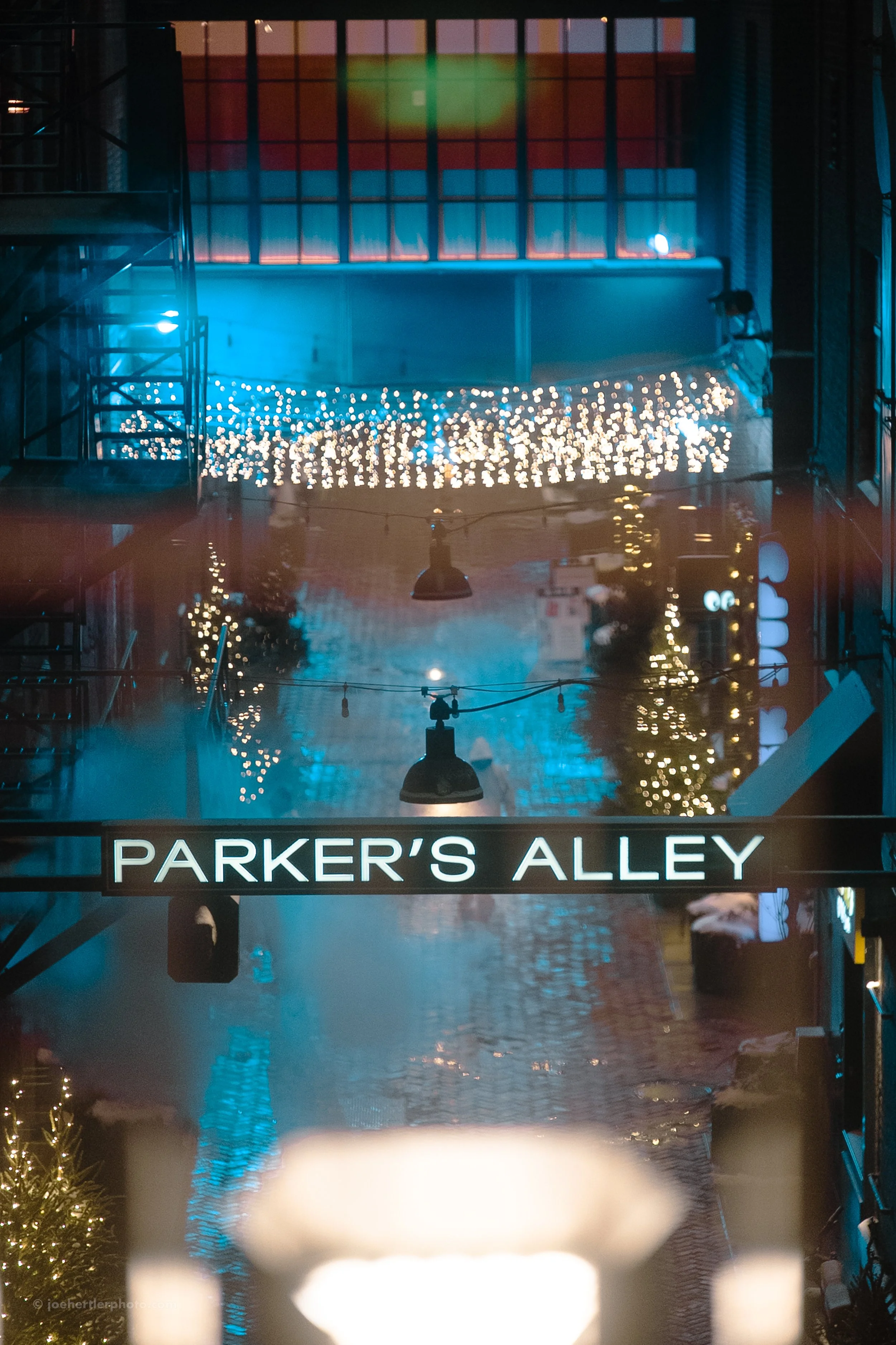 Nighttime scene of Parker's Alley decorated with string lights and Christmas lights reflected on the wet cobblestone street, with a neon sign reading 'PARKER'S ALLEY' hanging above.