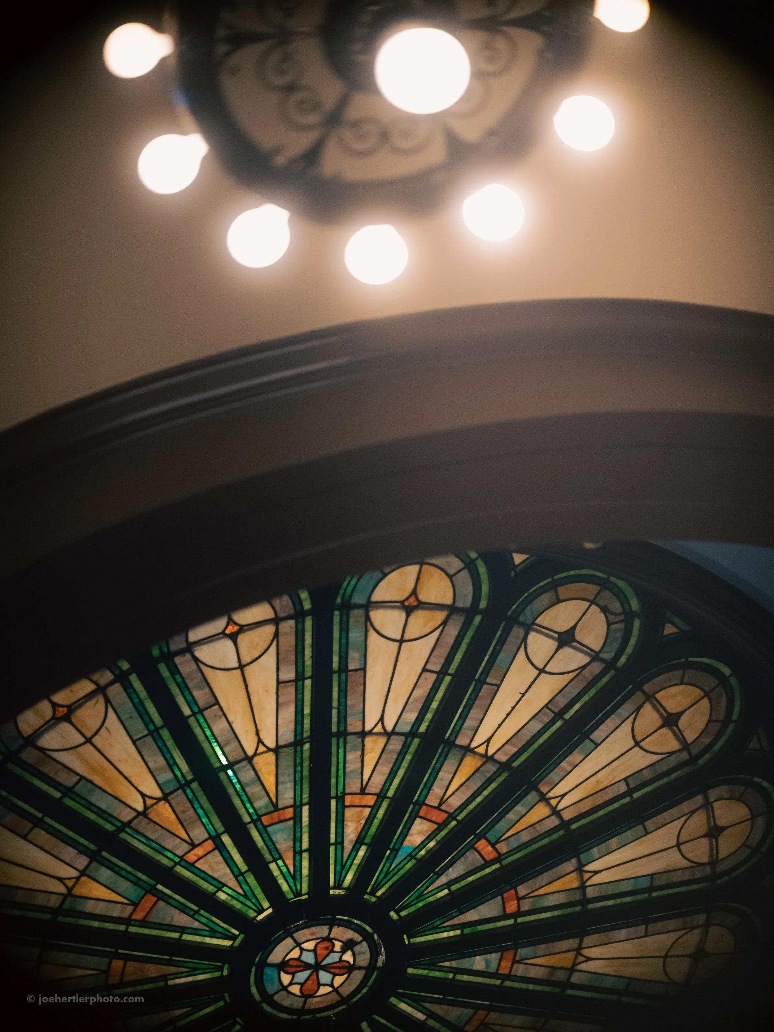 Looking up at a stained glass window with a circular design and geometric patterns, illuminated from behind. Above it, a chandelier with multiple round bulbs is also visible.