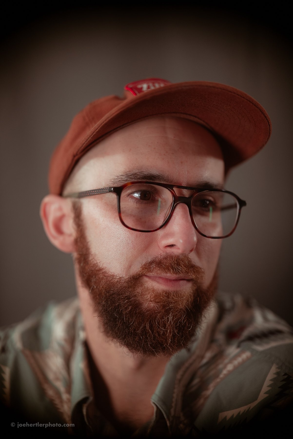 Close-up of a man with a beard and glasses wearing a brown cap. He is looking slightly to the side with a neutral expression.