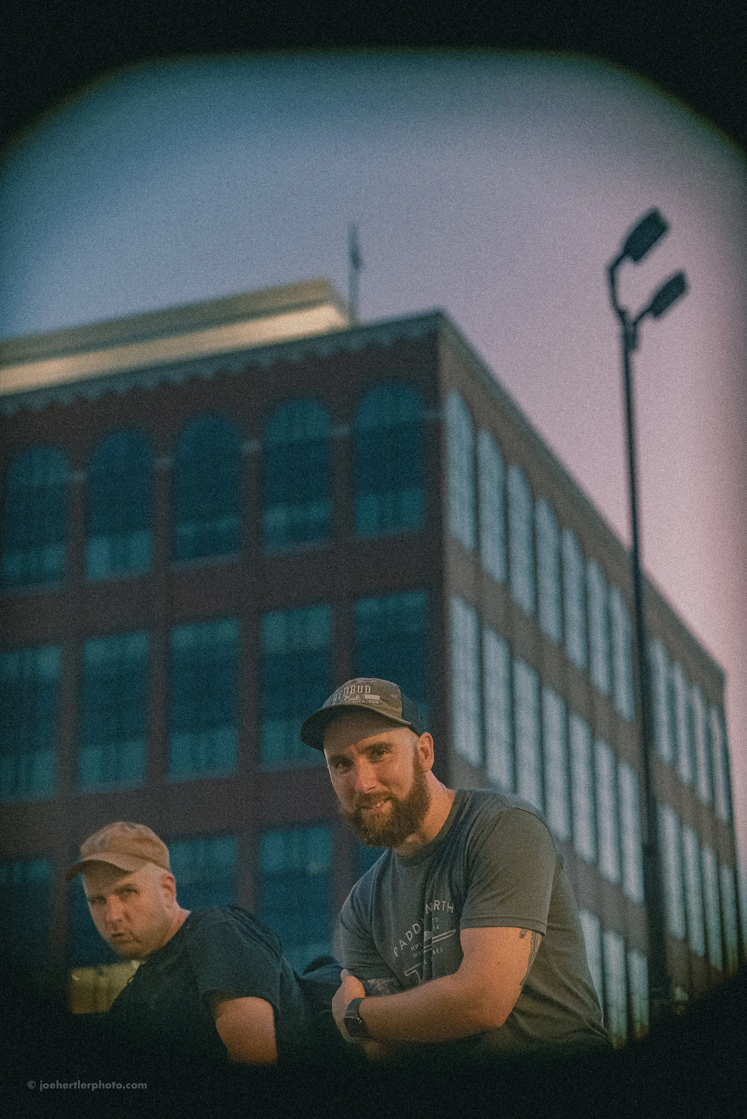 Two men sitting outdoors in front of a building during evening, with a large building and a streetlight in the background.