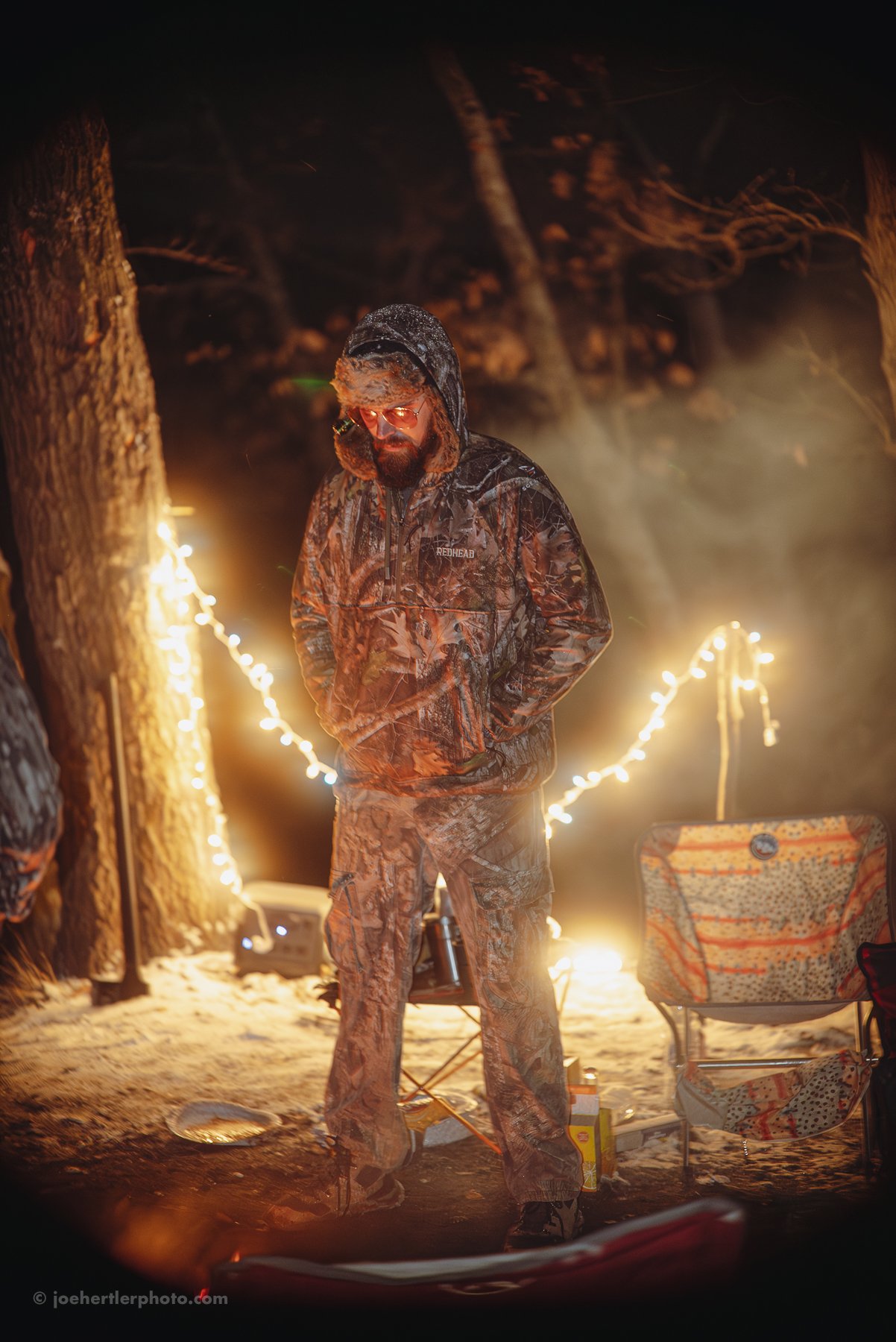 A person dressed in camouflage winter clothing, including a hooded jacket and pants, standing outdoors at night with string lights and equipment around him.