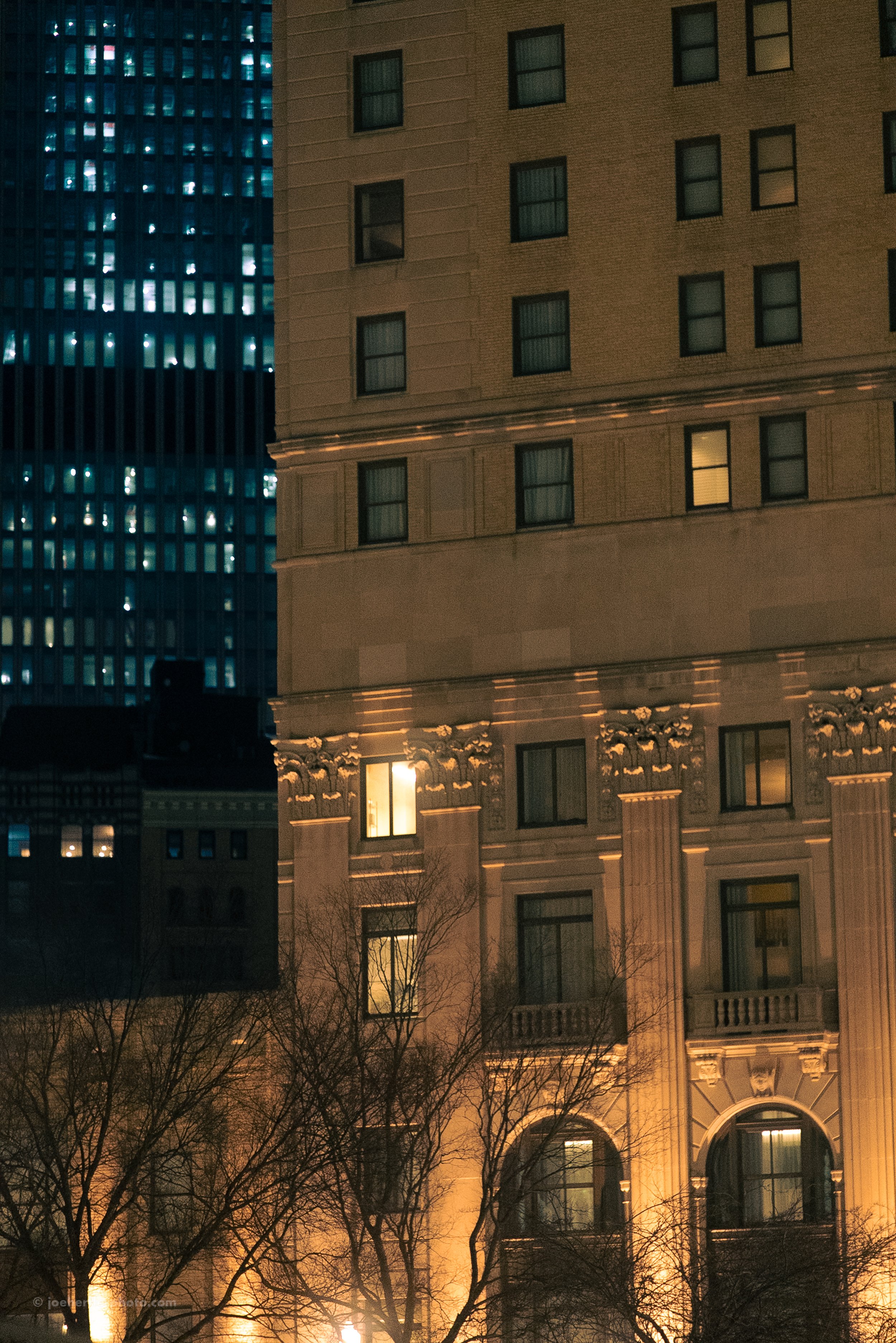 Nighttime cityscape with illuminated windows in tall buildings and leafless trees in the foreground.
