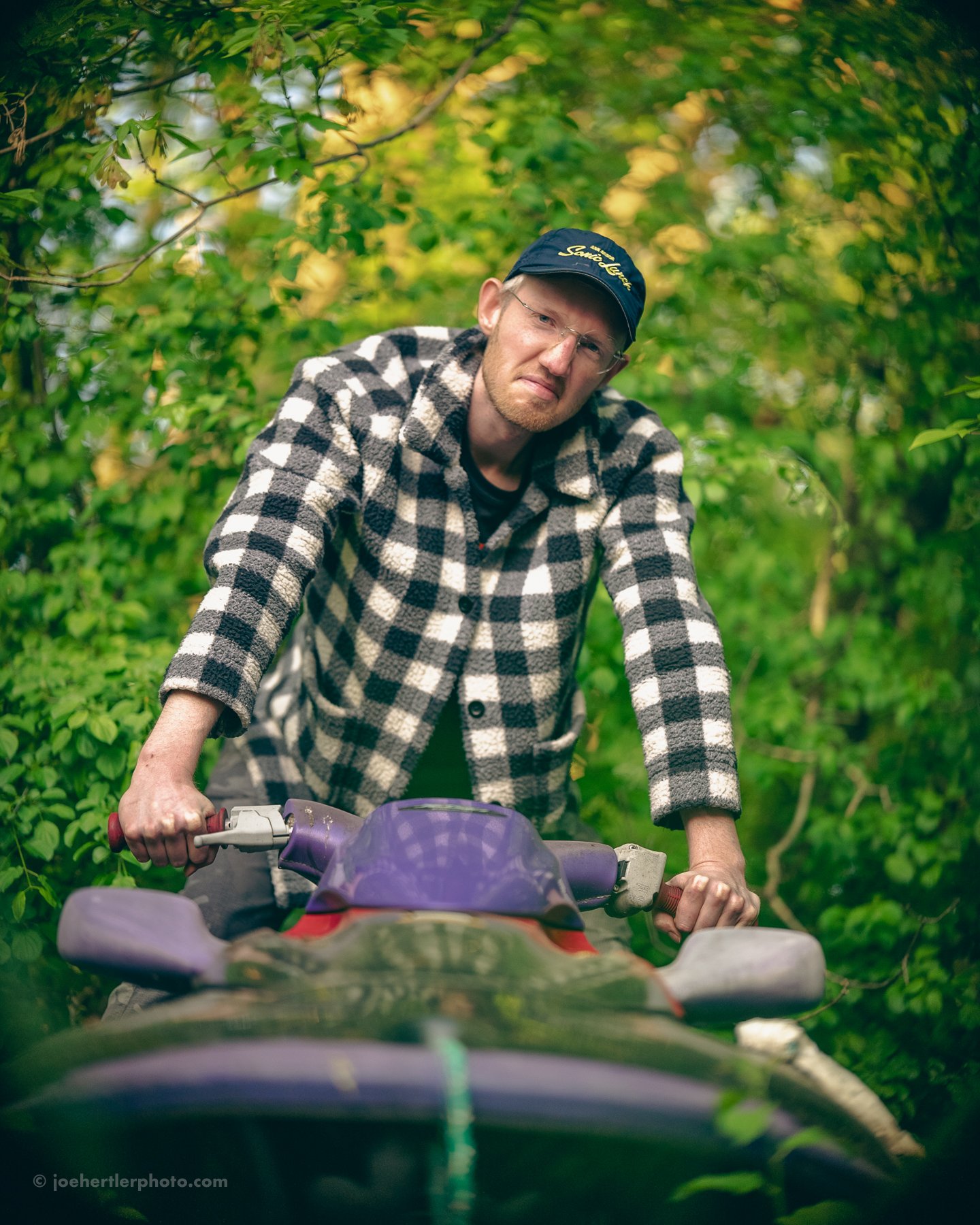 A man wearing a black cap and a black-and-white checkered jacket is riding an ATV through a lush green forest, with dense foliage surrounding him.
