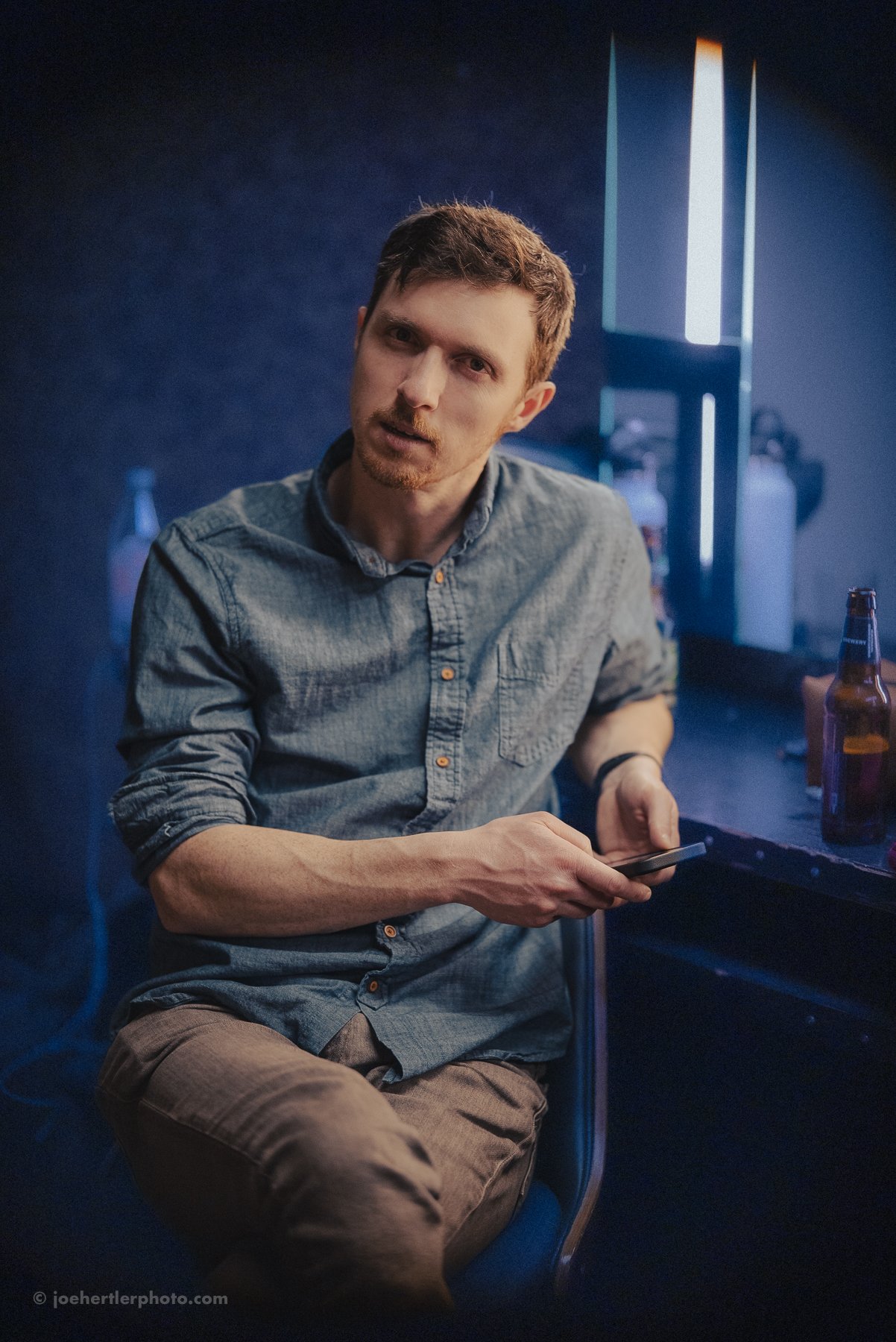 A young man with short brown hair and a beard, wearing a denim shirt, sitting in a dimly lit room with a blue background, holding a smartphone in his right hand, surrounded by bottles on a counter.