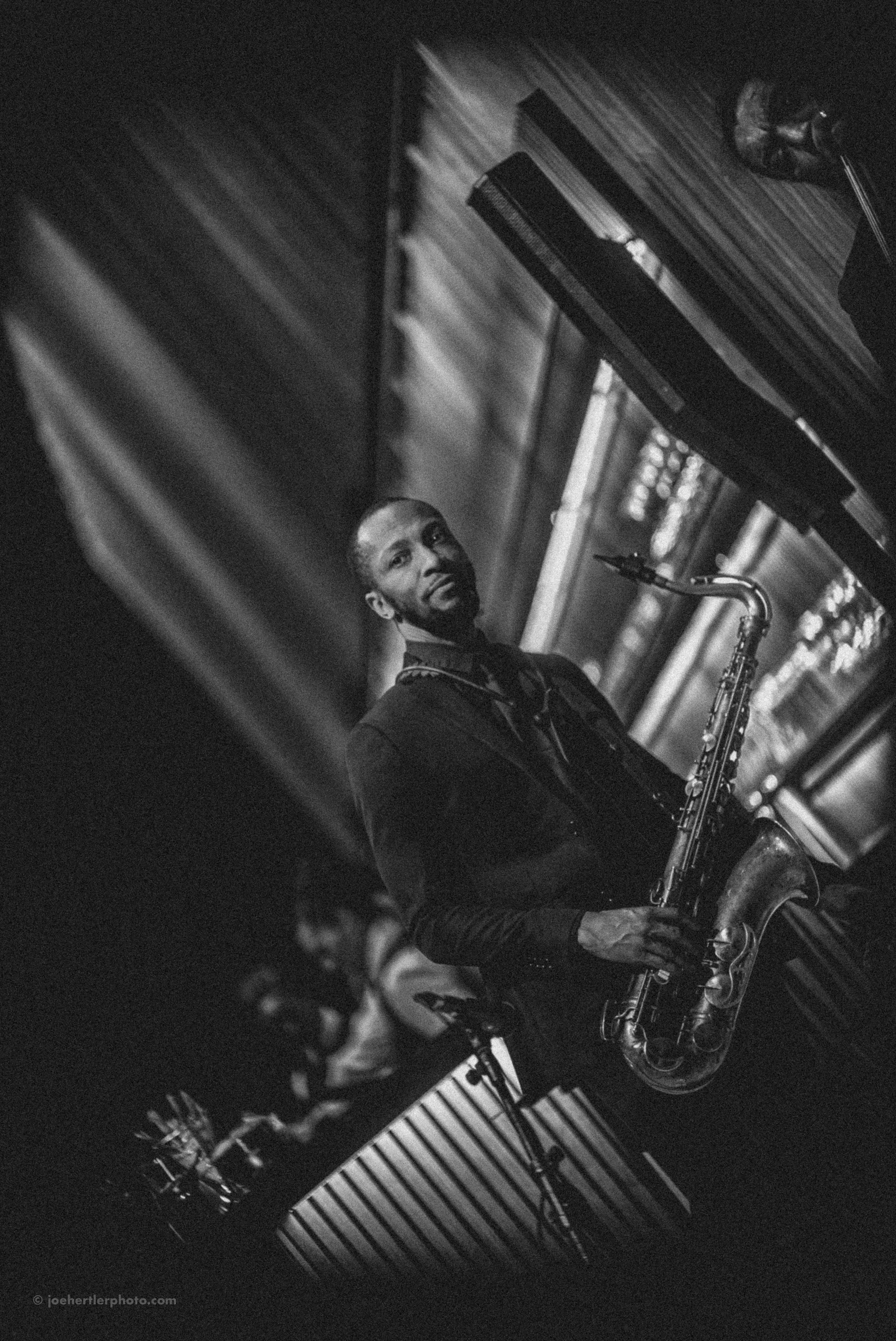 Black and white photo of a jazz musician playing a saxophone, standing near a piano, with wooden paneling in the background.