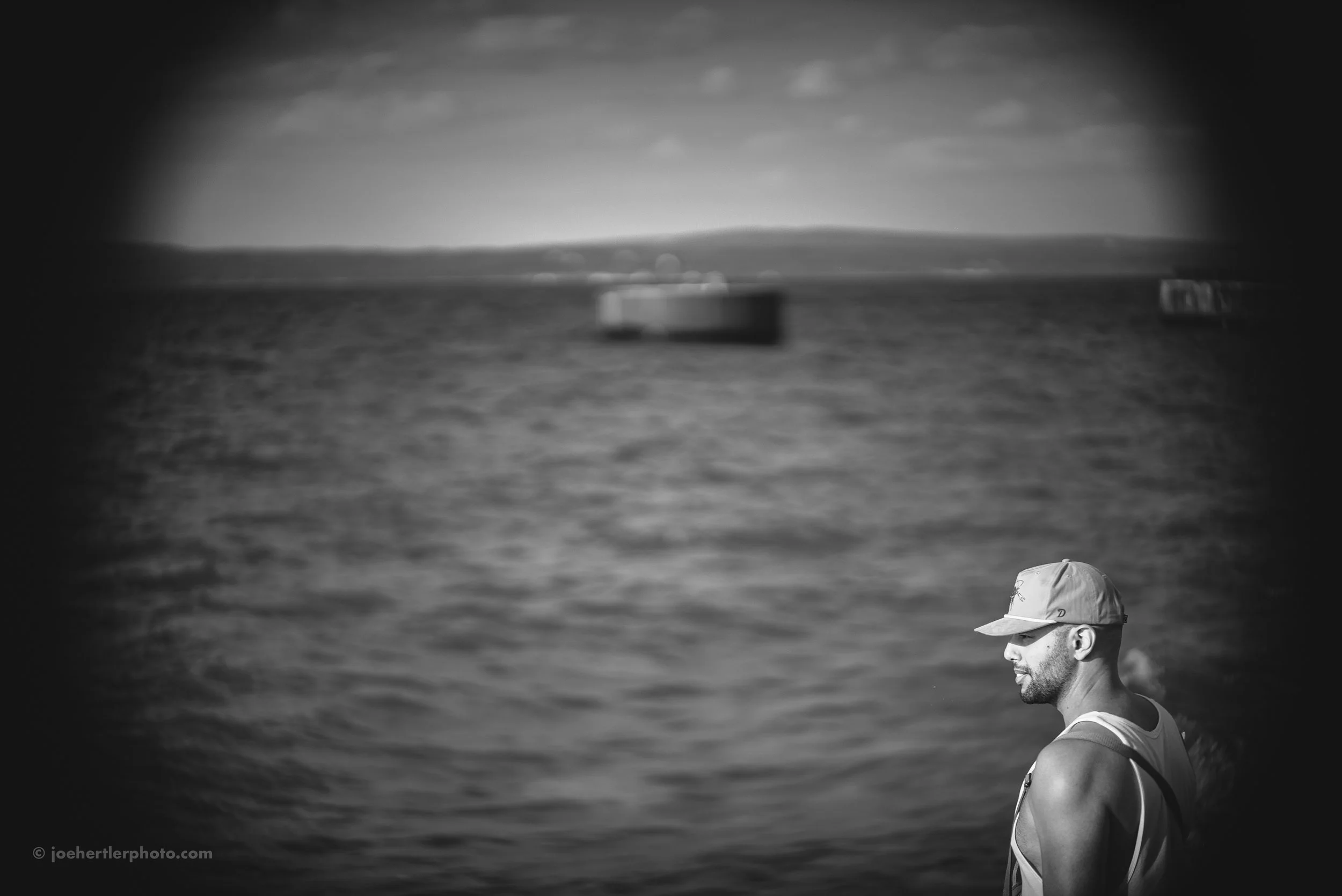 A black and white photo shows a man with a beard wearing a baseball cap and sleeveless shirt, standing on the water with a boat and distant shoreline in the background.