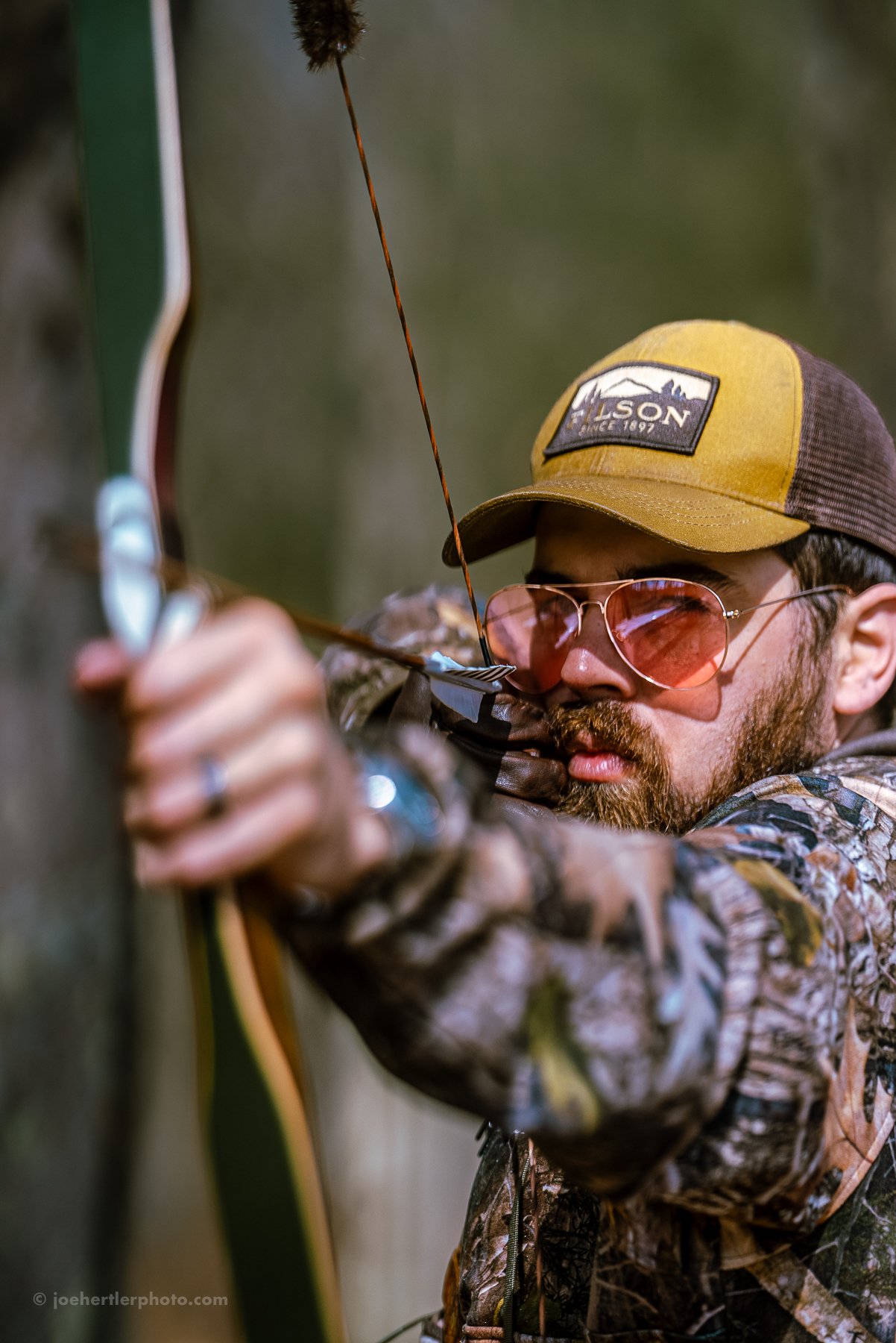A man aiming a bow and arrow, wearing a camouflage jacket, sunglasses, and a brown and yellow baseball cap.
