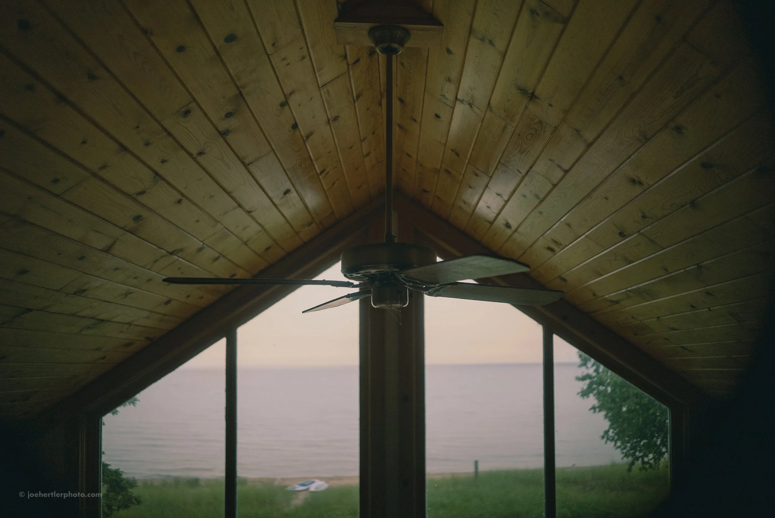 Interior of a room with a wooden vaulted ceiling, a ceiling fan in the center, and large windows showing a view of a lake or ocean and some greenery outside.