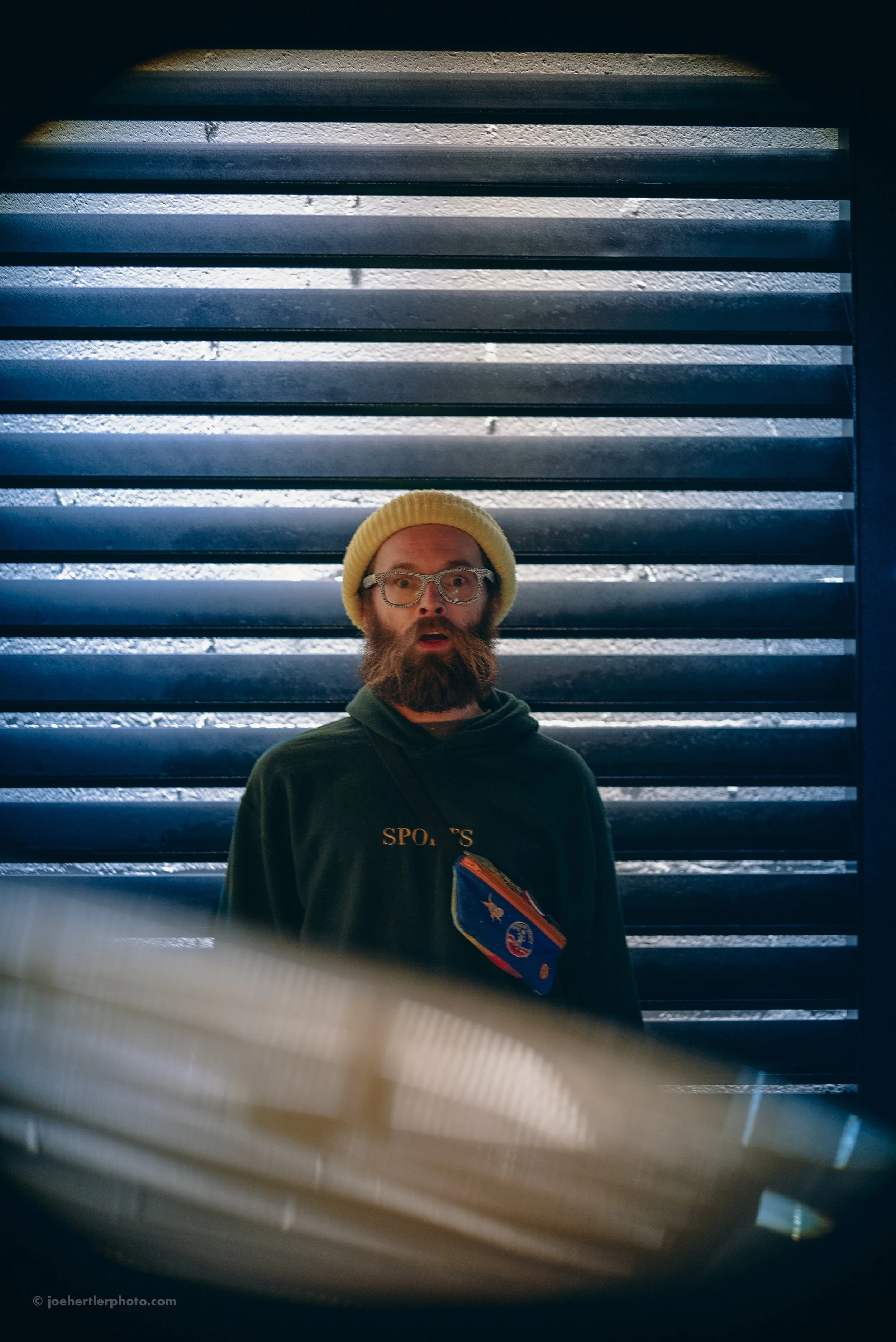 A man with glasses, a beard, and a yellow beanie stands in front of black horizontal blinds, with a surprised expression on his face.