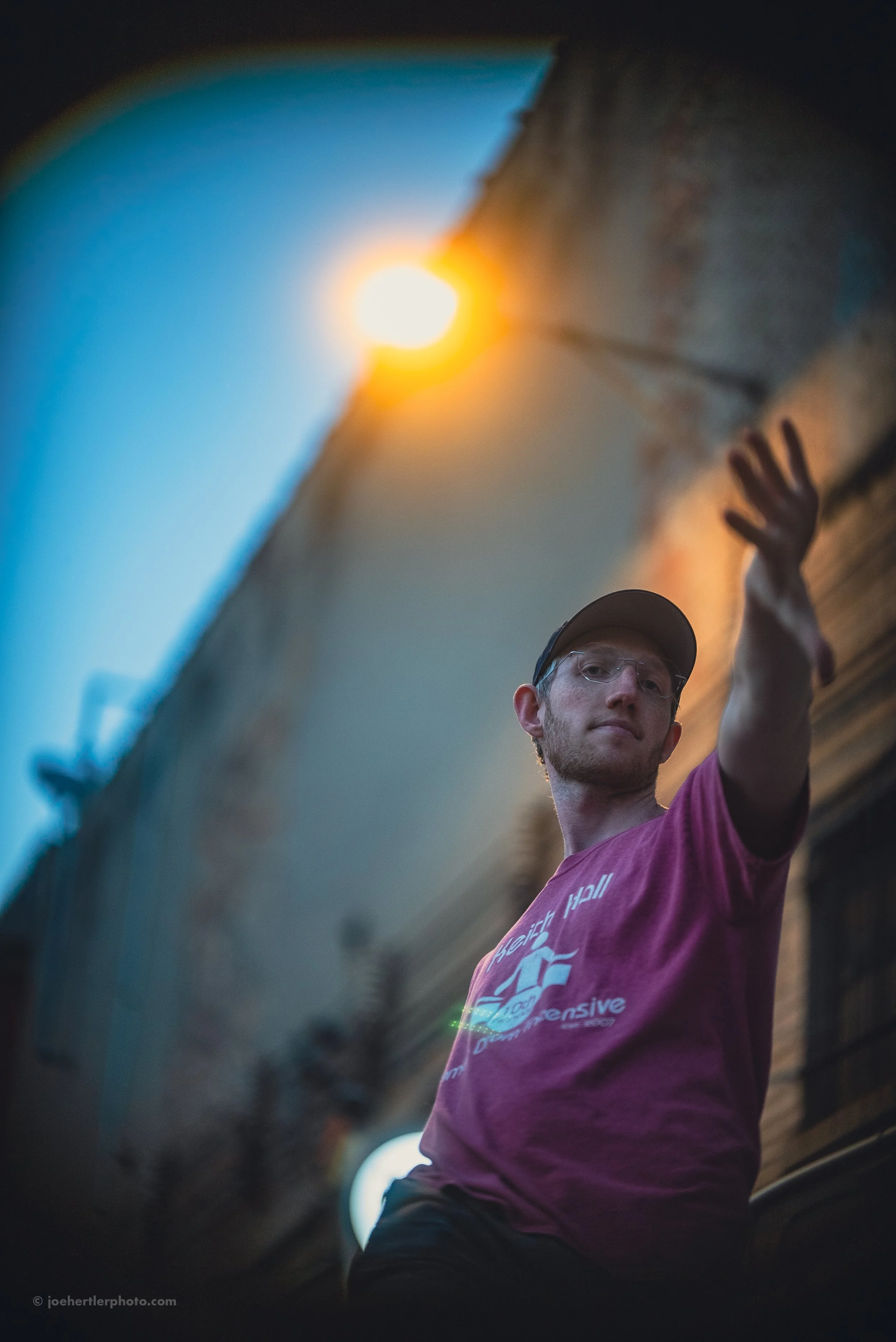 A young man wearing glasses, a cap, and a maroon T-shirt reaches upward with his right hand outdoors during sunset or dusk, with a building wall and a bright orange sun in the background.