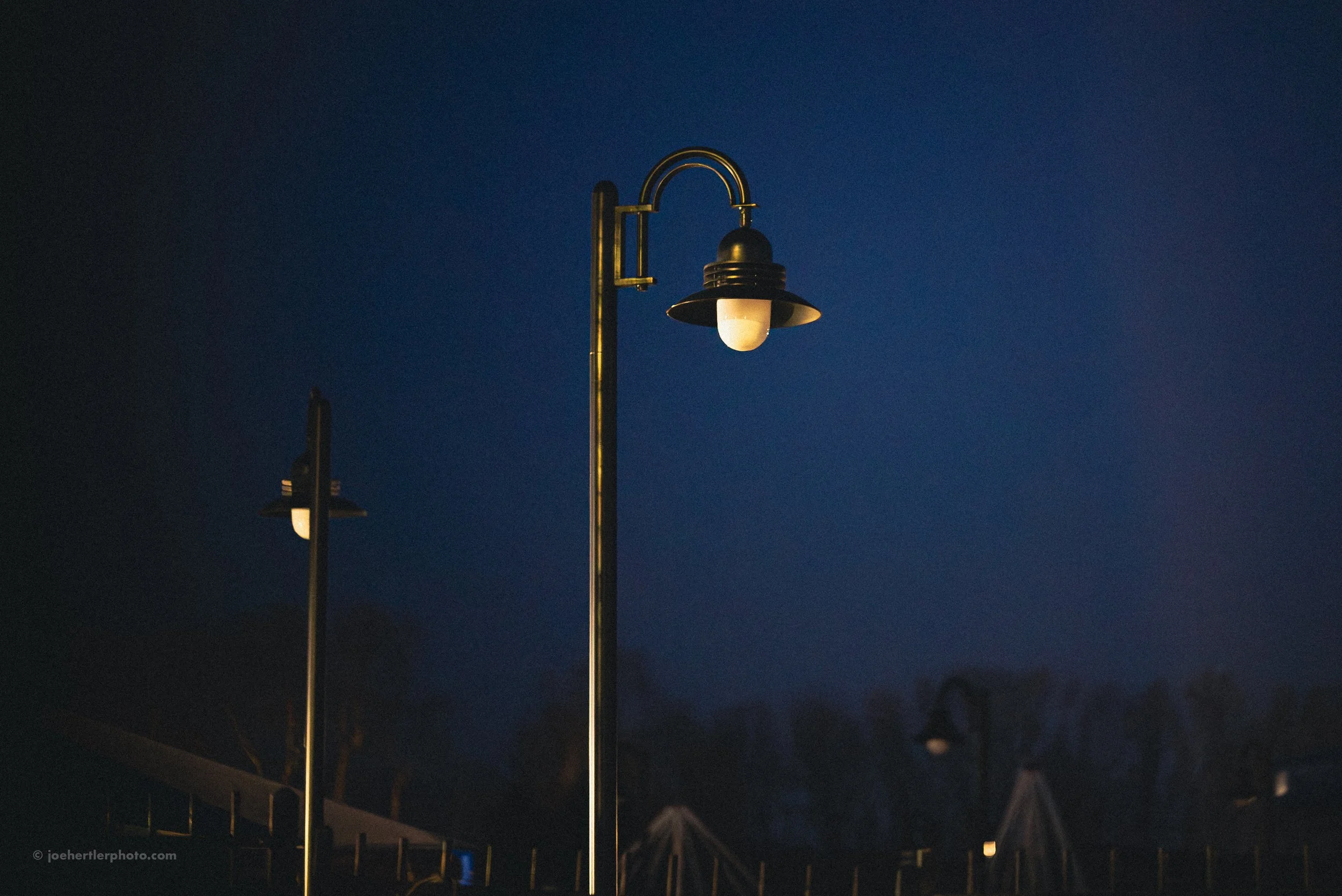 Two street lamps illuminated against a dark evening sky. The larger lamp is in focus, with a smaller one further in the background.