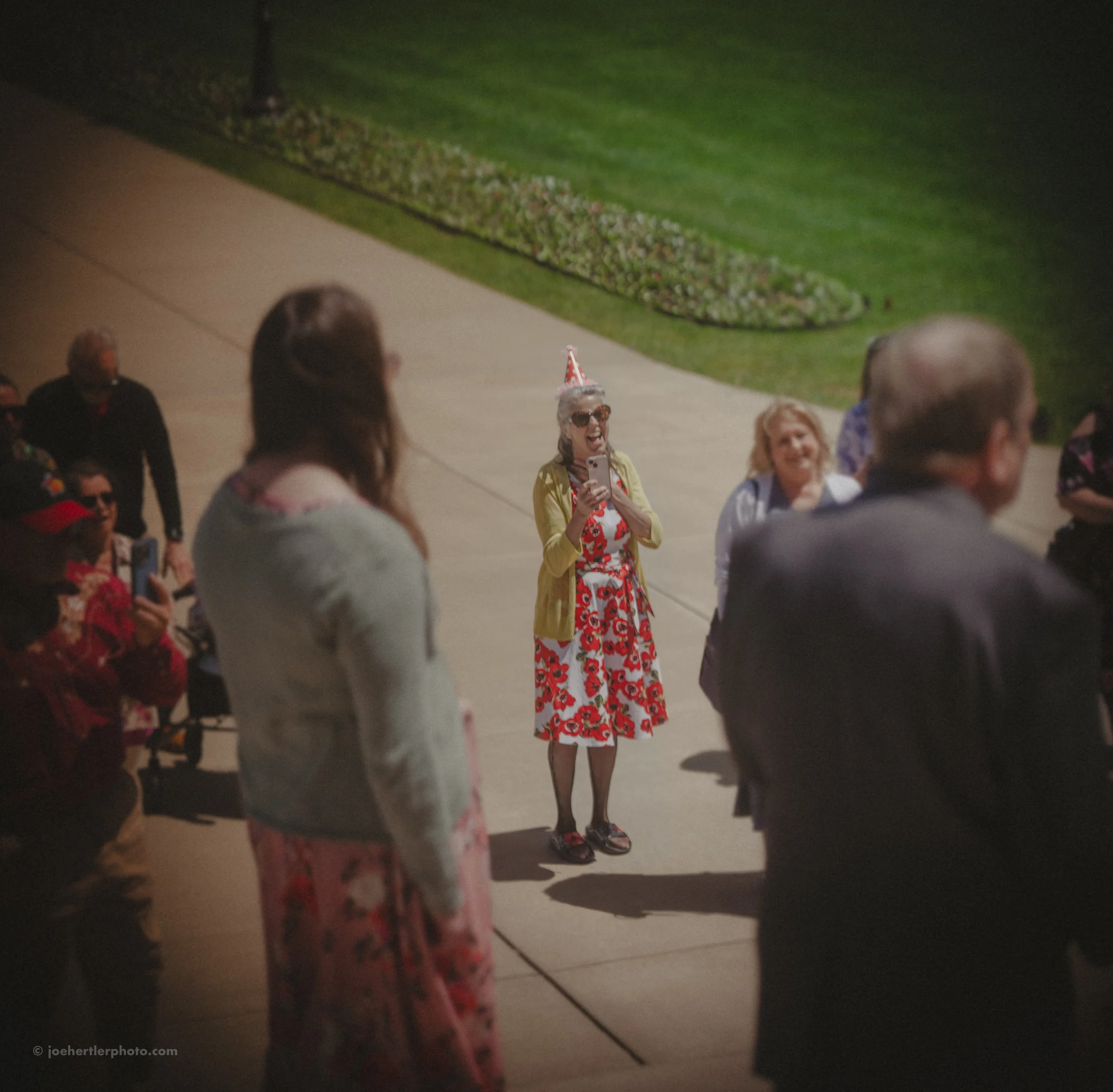 Woman wearing a red floral dress, yellow cardigan, sunglasses, and a party hat taking a selfie with a group of people in an outdoor setting.