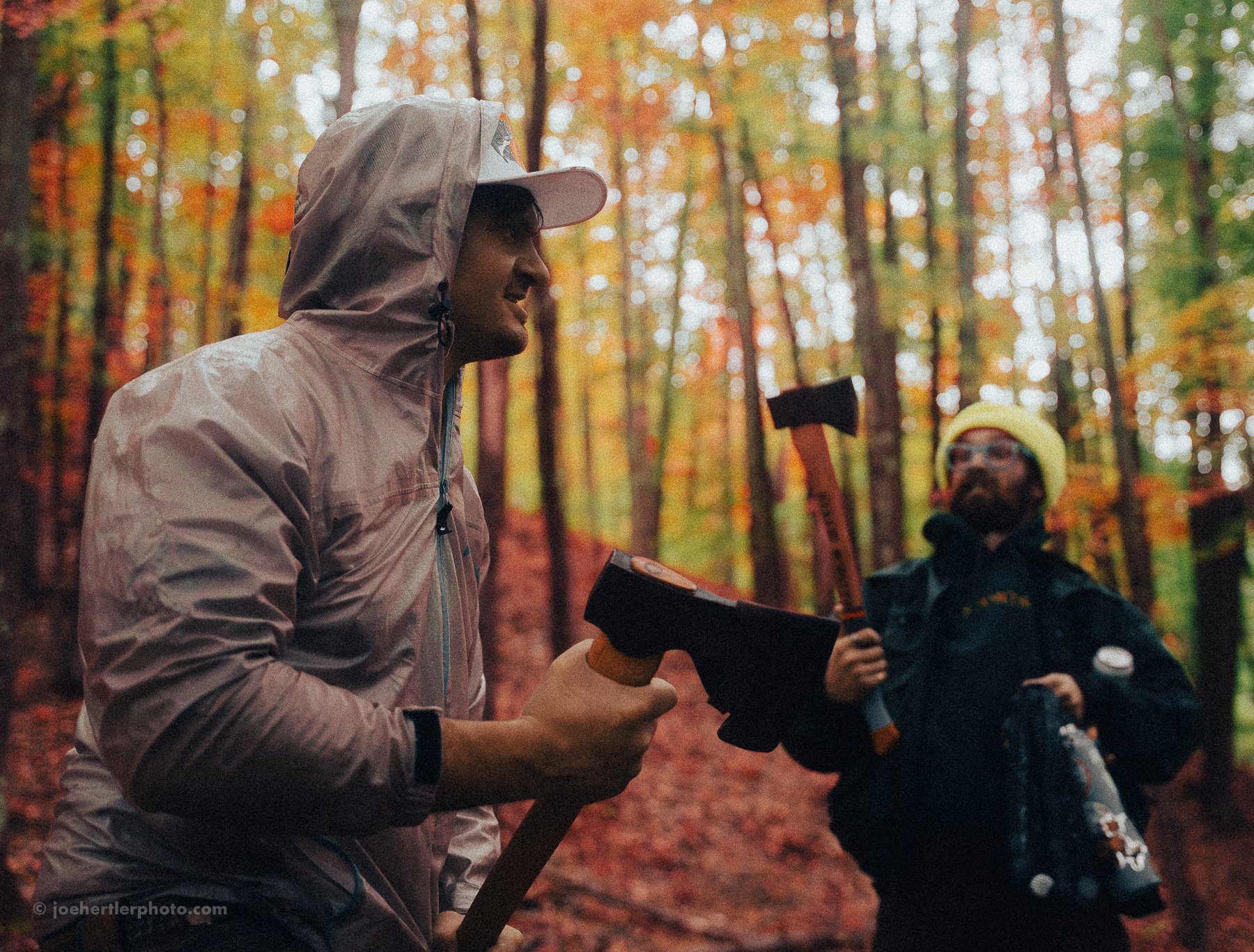 Two men standing in a forest during fall, one holding an axe and the other holding a chipper, enjoying a conversation.