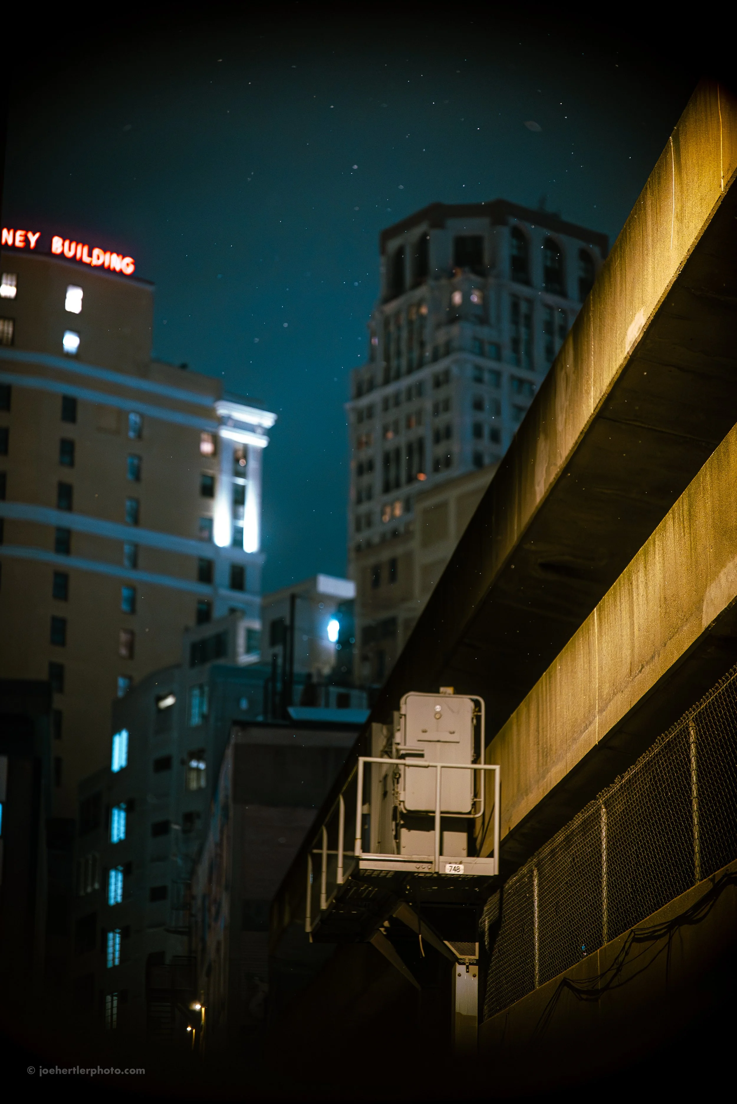 Nighttime cityscape with tall buildings, illuminated windows, and a construction platform with equipment in the foreground.