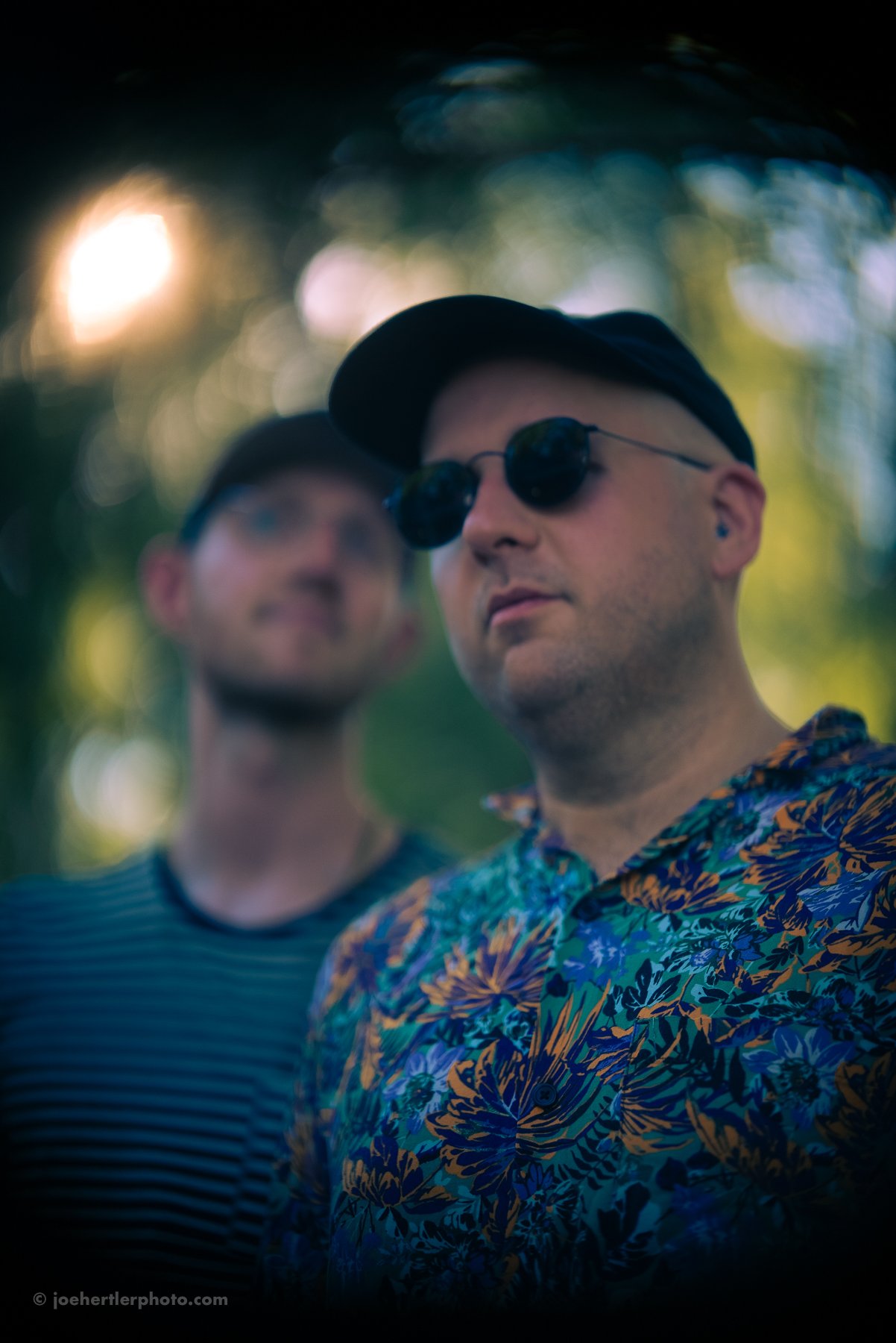 Two young men standing outdoors with one in focus wearing a floral shirt, sunglasses, and a hat, and the other slightly blurred behind him, both in a natural setting with sunlight filtering through trees.