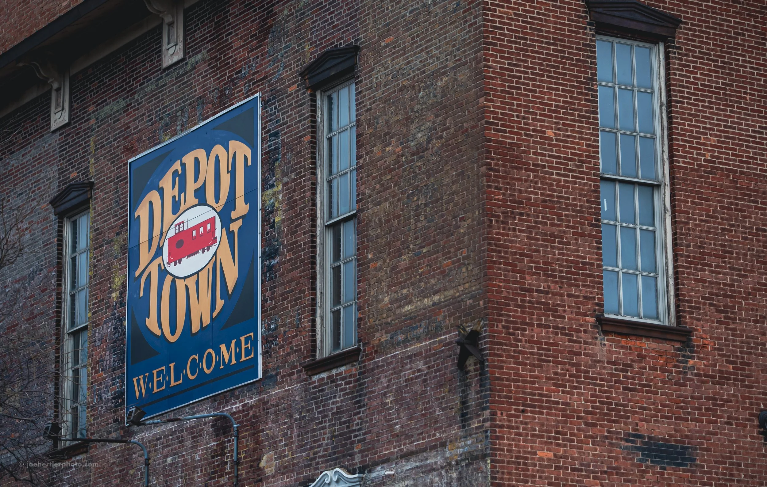 A brick building with three tall windows and a DePot Town sign that features a red vintage trolley and a welcome message.