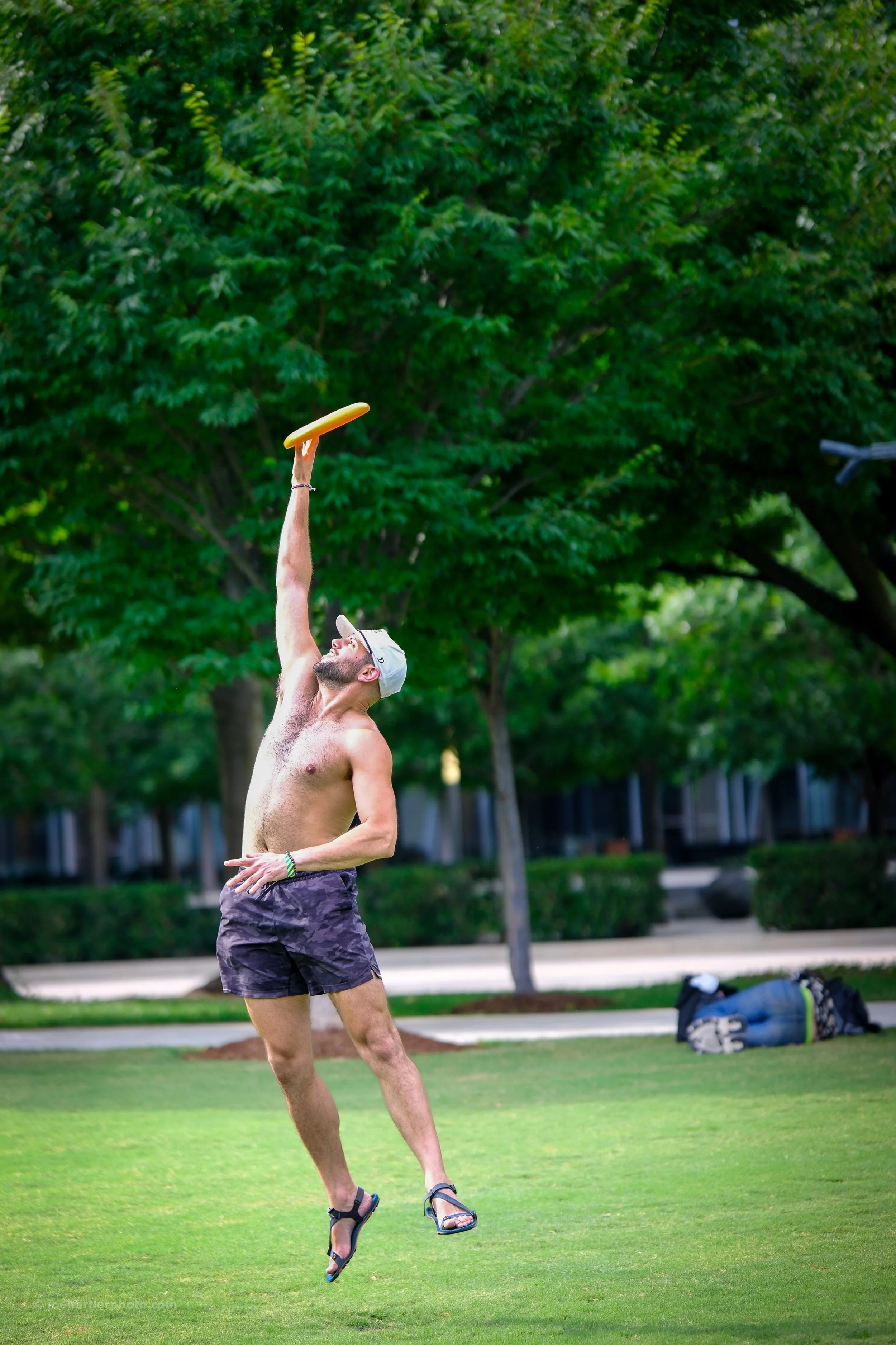 A shirtless man wearing a gray cap, shorts, and sandals is playing disc golf outdoors, reaching up to throw a yellow disc amidst green trees and a grassy field.