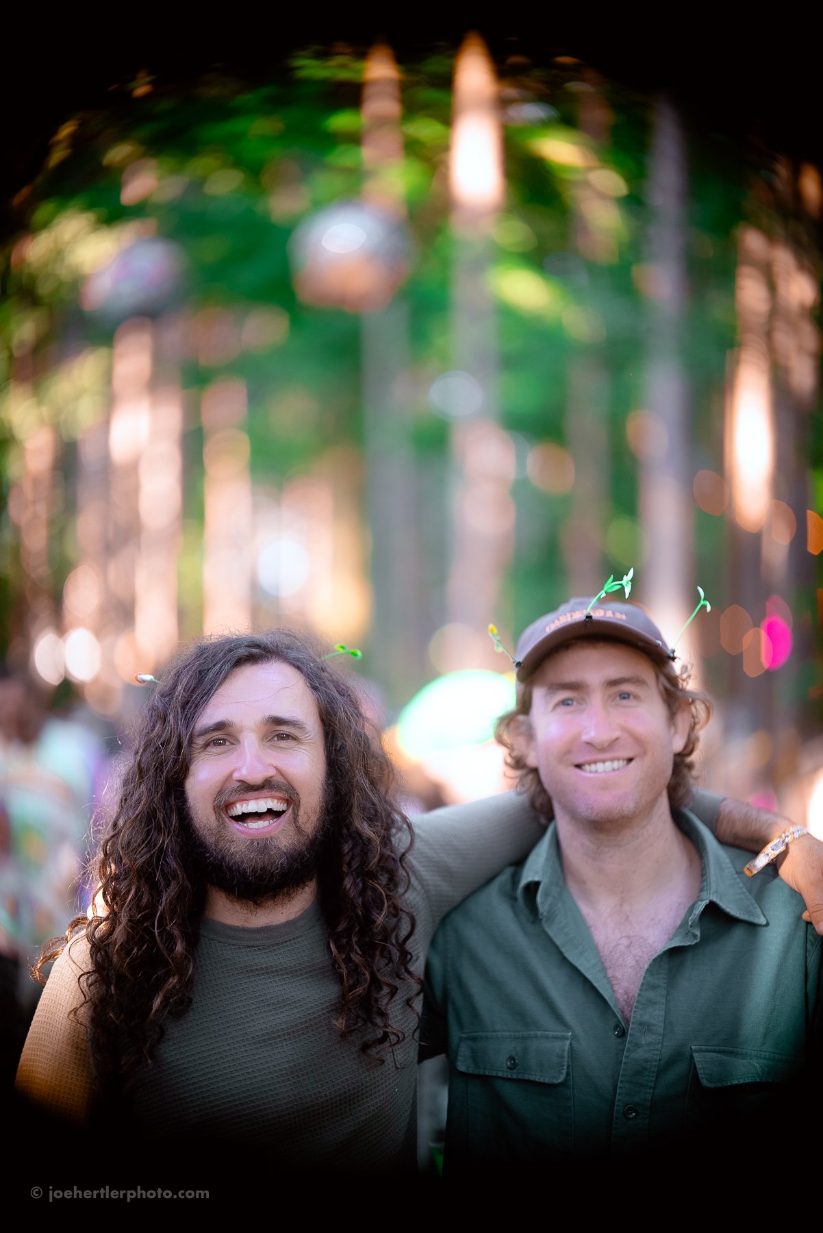 Two men smiling with their arms around each other at an outdoor celebration or festival, wearing casual clothing and novelty headwear, with a blurred colorful background of lights and trees.