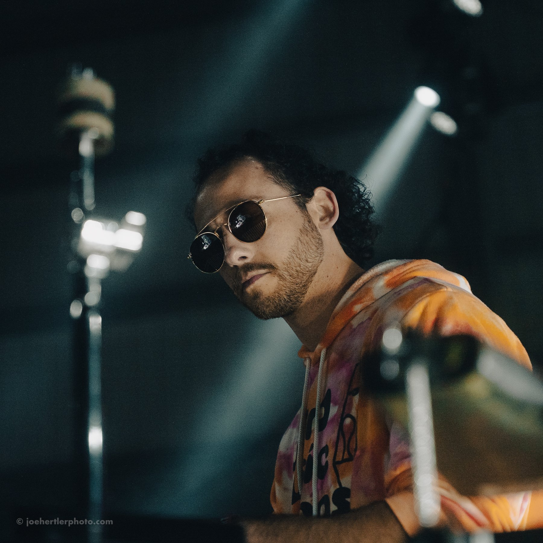 A man wearing sunglasses and a colorful hoodie playing the piano in a dimly lit setting.