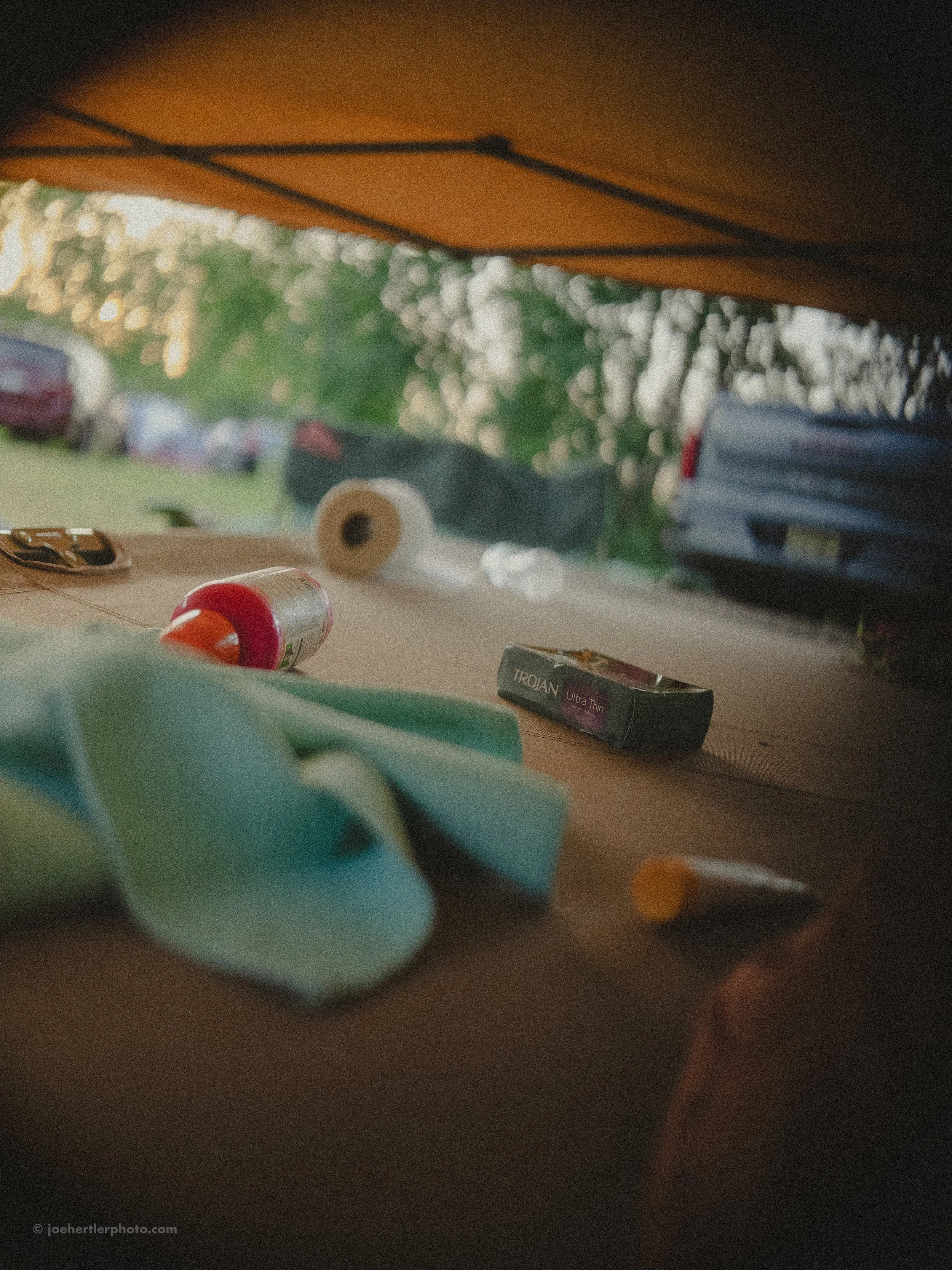 View from inside a tent showing camping supplies on the ground, including a roll of toilet paper, a box labeled Trojan Ultra Thin, a small tub of ointment, a yoga mat or cloth, and a yellow pill or capsule, with cars and trees visible outside in the 