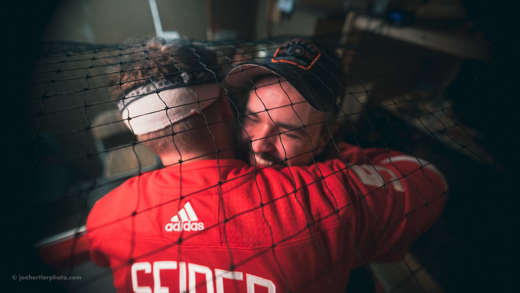 Two men in a sports setting embrace each other through a metal fence, with one smiling and the other facing away, wearing a red jacket with Adidas logo.