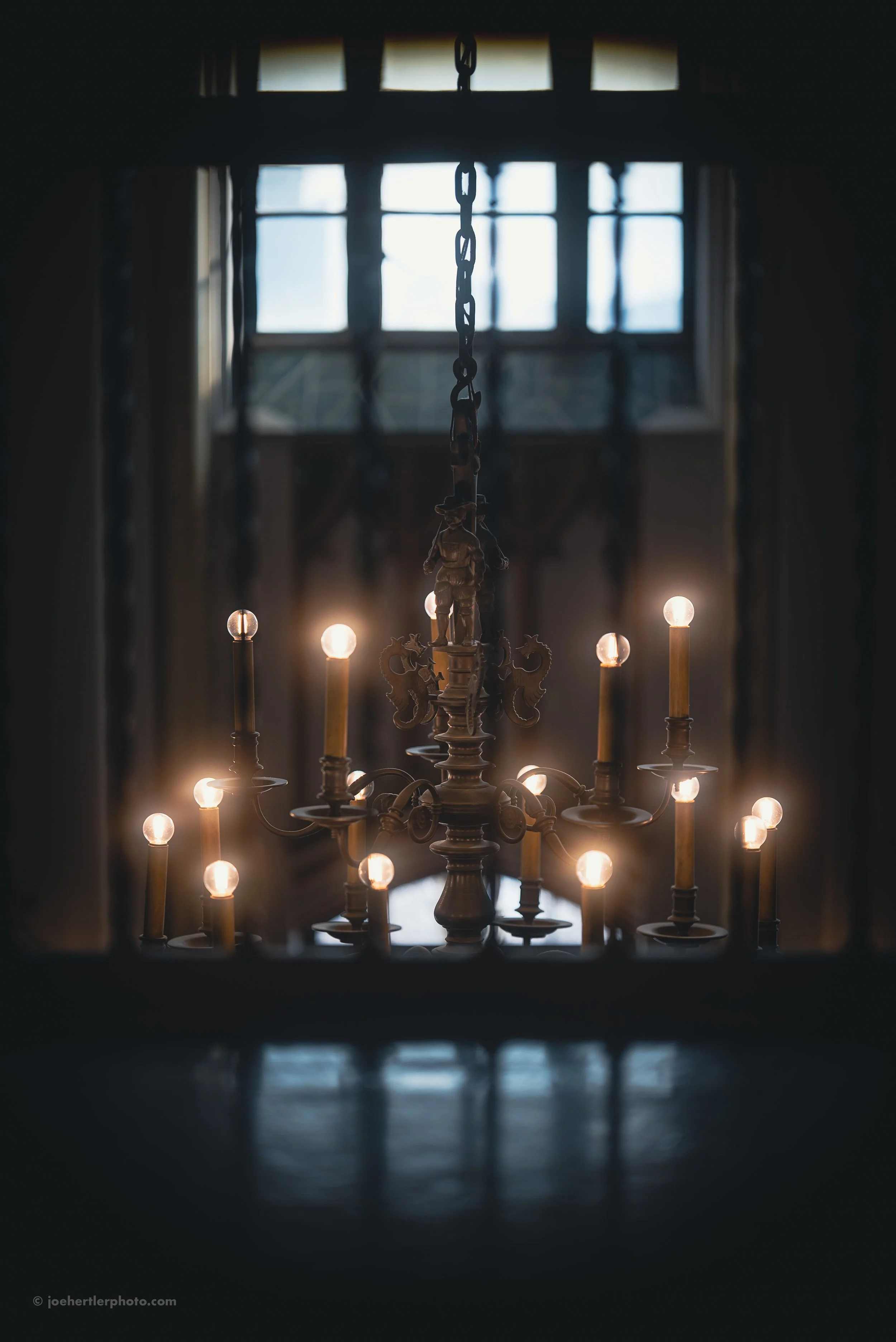 A chandelier with lit candles hanging from a chain, viewed through a window with multiple panes, in a dimly lit room.