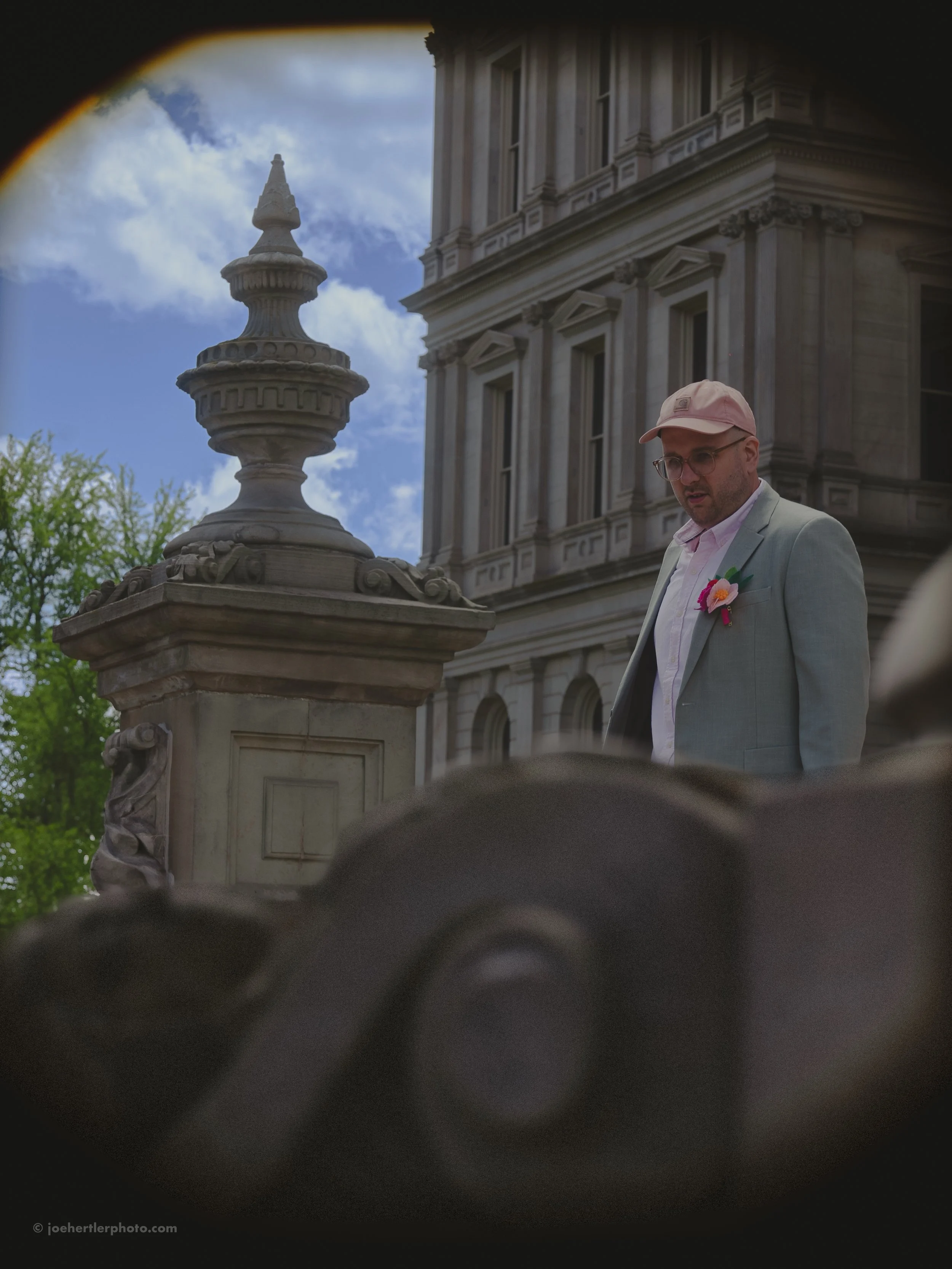A man wearing a light gray suit, pink shirt, pink cap, and glasses standing outside near an ornate stone railing and classic building with detailed architecture, under a partly cloudy sky.