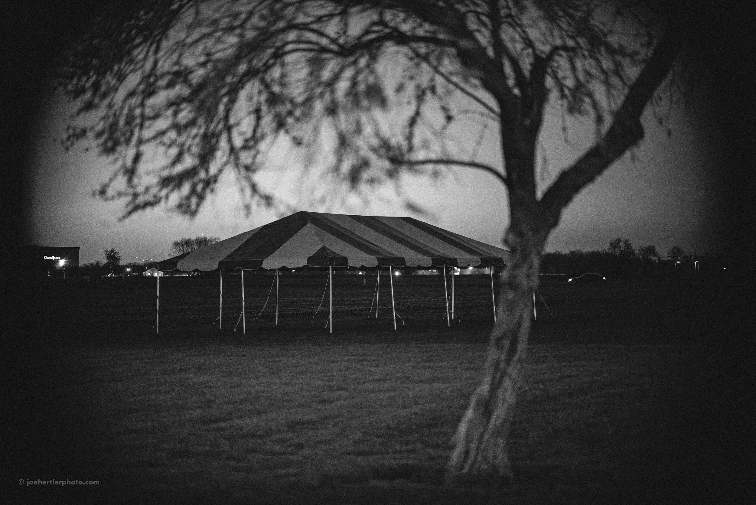 A nighttime outdoor scene featuring a large striped tent set up on a grassy field, with a leafless tree in the foreground and a few lights visible in the distance.